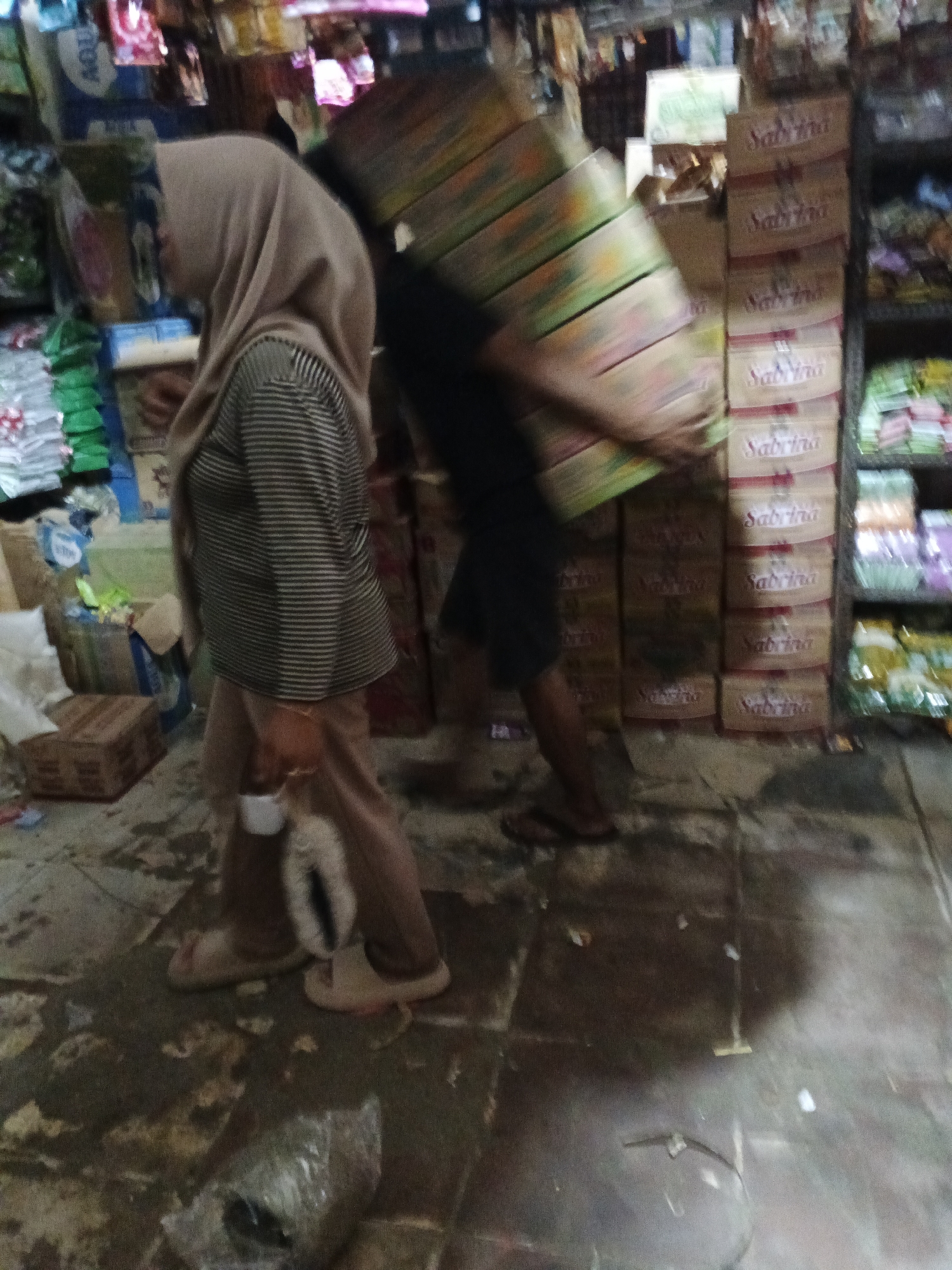 A low-angle, full shot captures a bustling street-level market scene in Warungkiara, Indonesia. A person wearing a light brown hijab and a striped shirt walks from left to right, carrying a white plastic bag. To their right, another person, partially obscured, walks away from the camera carrying a large stack of boxed goods on their left shoulder, the boxes are colorful and appear to be food items, possibly instant noodles. The ground is concrete and appears wet and dirty, with scattered debris. Shelves laden with various products, including boxes labeled "Sabrina," are visible in the background, suggesting a grocery or convenience store setting. The lighting is dim, creating shadows and a somewhat hazy atmosphere.