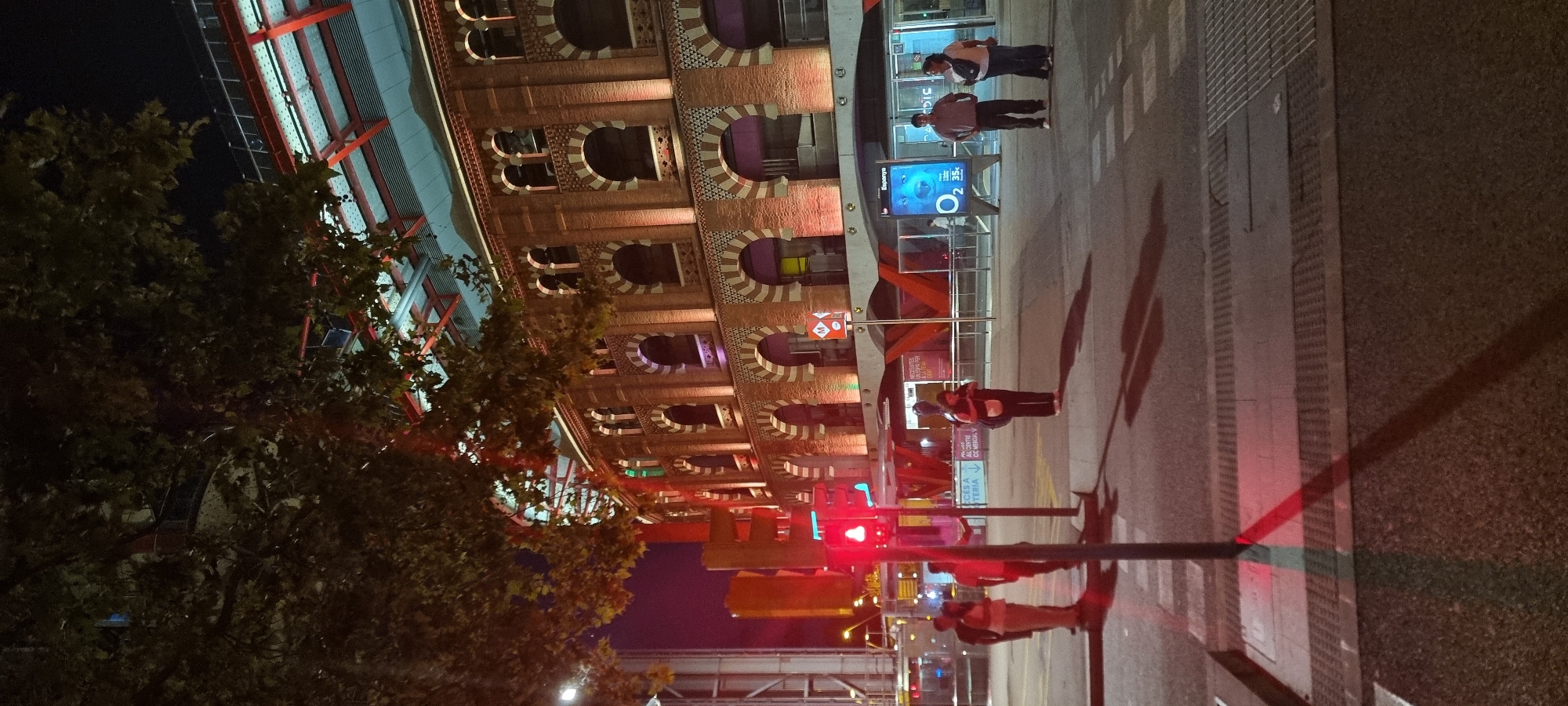 The image depicts a nighttime scene on a street in Barcelona, Spain. In the background, the ornate brick facade of a large building with arched windows is illuminated by warm lighting, suggesting it is a historic or significant structure. The building appears to be the Las Arenas bullring, now a shopping center, known for its distinctive architecture. In the foreground, a red traffic light is visible, casting a red glow onto the wet asphalt. Several people are present, some standing at the crosswalk, waiting to cross the street. One woman is standing alone, facing the camera with her back to the building, carrying a large bag over her shoulder. To the left, a group of three individuals are gathered. The overall atmosphere is urban and nocturnal, with the artificial lights creating dramatic shadows.