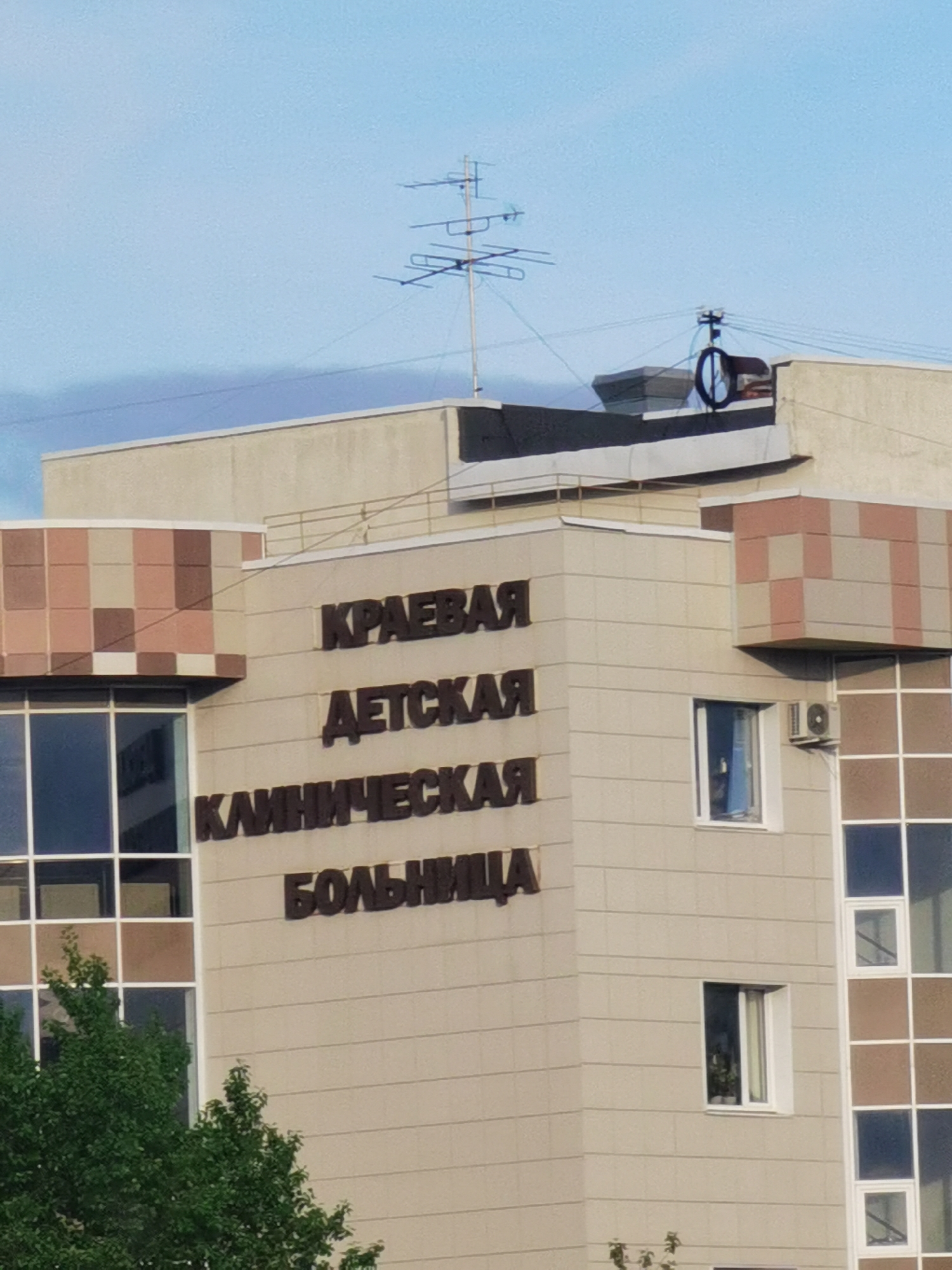 The image shows a section of a building identified by the brown lettering as "KPAEBAЯ ДETCKAЯ KЛИHИЧECKAЯ БOЛЬHИЦA" which translates to "Regional Children's Clinical Hospital." A Google search suggests this is located in Perm, Russia. The building is multi-story with beige exterior cladding and features checkered brown accent pieces on its corners. Some windows are visible. An air conditioning unit is mounted beneath one window. A large TV antenna stands on the roof. Trees partially obscure the building's lower portion. The sky is blue with a few clouds.