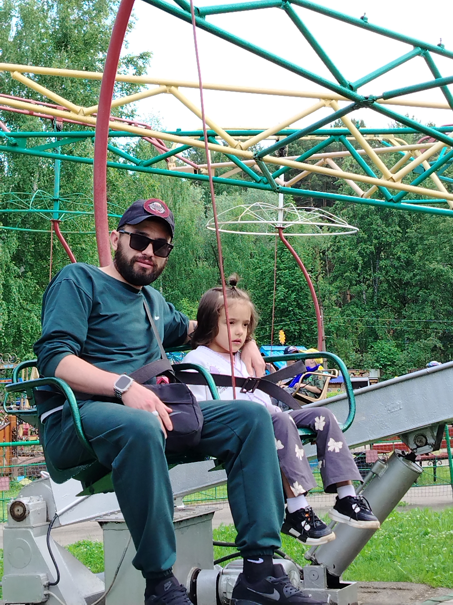 An outdoor image captures a man and a young child seated side-by-side on a green metal gondola-style amusement park ride in what appears to be an amusement park in Chaikovsky, Russia.
The man, positioned on the left, has a dark beard and wears sunglasses, a black baseball cap with an unidentifiable red and white patch, a dark green long-sleeved shirt with a white Nike swoosh, and matching dark green pants. He has a silver-colored watch on his left wrist and a black shoulder bag with a white Nike swoosh resting on his lap. He looks directly at the camera with a calm expression.
To his right, a young child, likely a girl, with dark hair pulled back in a small bun, is seated. She wears a white t-shirt and gray pants adorned with white butterfly or bow patterns, and black and white sneakers. She is looking towards the right side of the frame, away from the camera, appearing calm or pensive. Both individuals are secured by black safety harnesses across their laps.
The ride's structure features prominent red, green, and yellow metal beams forming a complex overhead lattice. In the background, lush green trees and foliage suggest a natural, wooded setting. The sky is bright, indicating daylight, possibly an overcast day. The ground below the ride appears to be grass. The image conveys a leisurely moment at an amusement park.