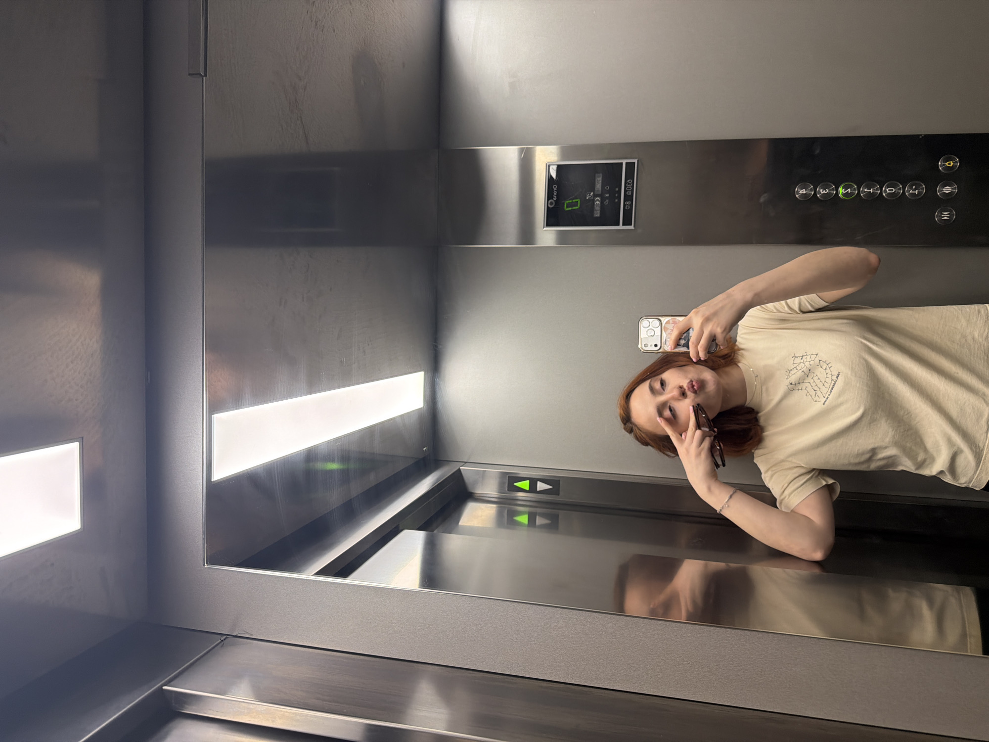 A young woman with auburn hair is taking a mirror selfie in an elevator. She is wearing a light-colored t-shirt and has her right hand held up to her face, making a peace sign with two fingers. She is holding her phone in her left hand to take the picture, and a pair of sunglasses are perched on her fingers. The elevator has stainless steel walls and ceiling, with bright white lights. There are elevator buttons and a digital display visible on the right-hand wall. The woman's reflection is visible in the elevator doors on the left. The setting is a modern elevator. The image does not contain any information that would allow for its location in Strasbourg, France to be determined.
