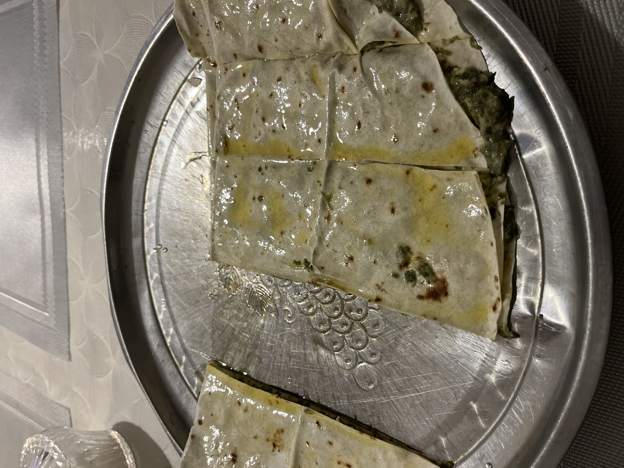 A high-angle, close-up shot shows a metal serving tray containing cut portions of a layered pastry dish. The pastry appears to have a spinach filling and is glistening with what appears to be oil. The tray is circular and features a decorative embossed design in its center. The background includes a white, textured tablecloth. Part of a clear glass object is visible at the bottom left. The photo may have been taken indoors, possibly in Kaspiysk, Russia.
