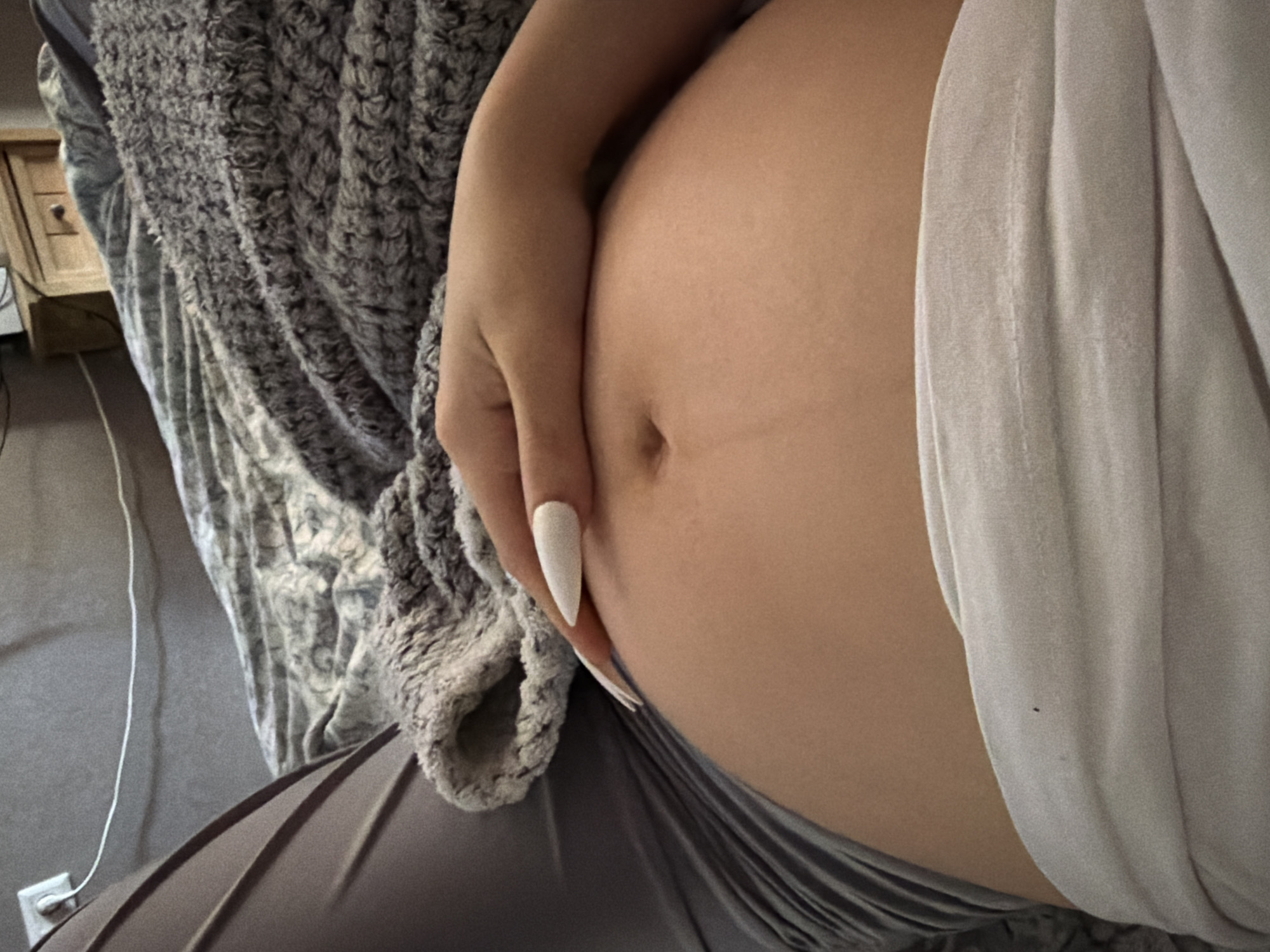 The image shows a close-up of a pregnant person's belly. Their hand, adorned with long, white pointed nails, rests gently on their stomach. They are covered by a textured, gray blanket and a light-colored piece of fabric, possibly clothing. The background features a hint of a wooden object with a drawer and knob, and a white electrical cord running to an outlet on a grey wall. The scene appears to be indoors, likely in a home setting. There is no discernible time of day or weather information. The focus is on the intimate portrayal of pregnancy. No text is visible.