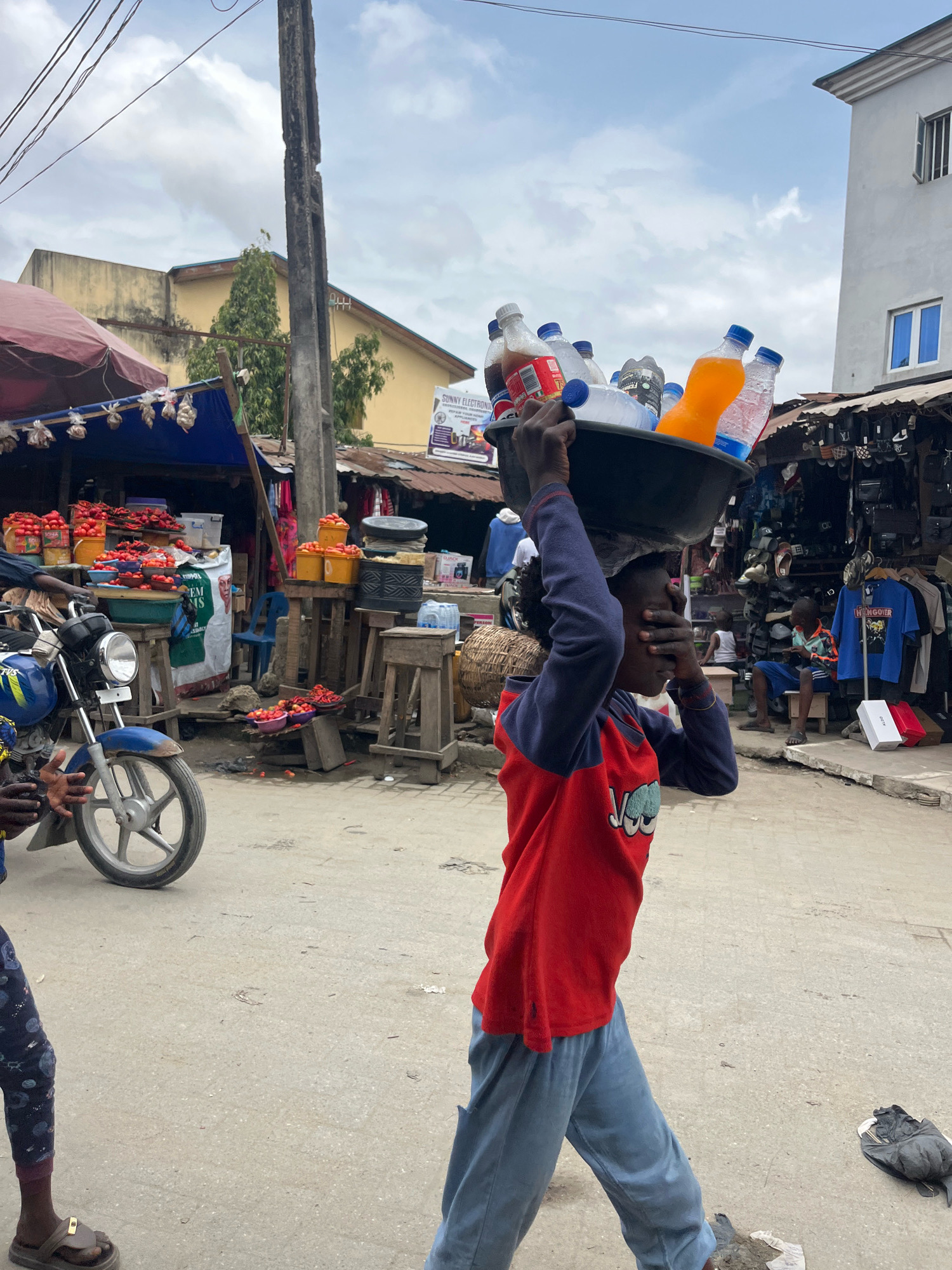 In Ajah, Nigeria, a young person carries a large black bowl filled with plastic bottles of various beverages on their head. The person wears a red long-sleeved shirt with blue accents on the sleeves and light blue pants. They are walking on a dusty, paved road in what appears to be a market area. To the left, a blue motorcycle is parked, and stalls with red tomatoes and other produce are visible. In the background, there are more market stalls selling clothing and other items, along with buildings and a utility pole. The sky is partly cloudy.