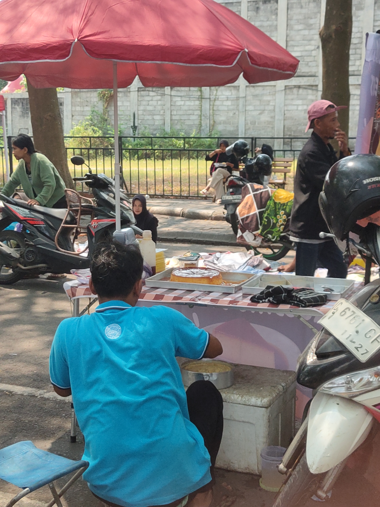 A street food vendor is preparing food at a market stall in Ngasem, Indonesia, on a sunny day. The vendor, wearing a blue t-shirt, is seen from behind, focused on a silver pan filled with what appears to be a baked good, possibly a cake or pastry. The stall is covered by a large red umbrella, casting a shadow over the scene. To the right of the vendor, a white motorcycle is parked, its front fender and license plate visible. In the background, several other people are present, some standing near motorcycles and others sitting. A woman in a green jacket is seen near a parked motorcycle on the left, and another woman in a black hijab is observing from behind the stall. The ground is paved with asphalt, and a grey brick wall and some trees are visible in the distance. The overall impression is of a bustling outdoor market environment.