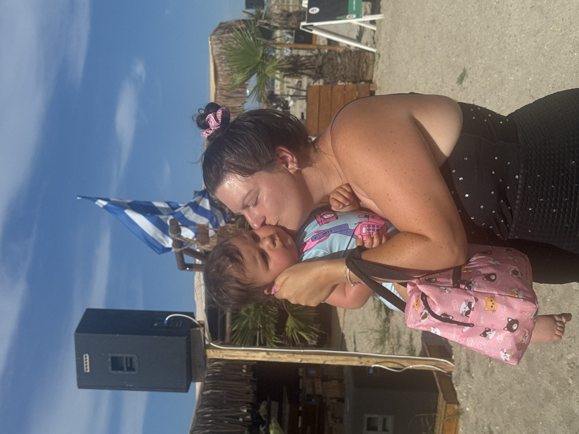 A woman, with her hair in a bun secured by a pink and white striped scrunchie, is kissing a baby on the cheek. The baby, wearing a light blue onesie with a pink camera and walkie-talkie print, is held in the woman's arms. A pink fabric bag adorned with cartoon cats is slung over the woman's shoulder, with the baby's foot visible protruding from it. The woman is wearing a black strapless top with silver embellishments. They are on a sandy beach, indicated by the ground texture and the presence of palm trees and a thatched-roof structure in the background. A large black speaker is mounted on a wooden structure to the left, and a Greek flag with blue and white stripes waves in the sky behind them. The scene appears to be a casual outdoor setting, likely a beach resort or event space.