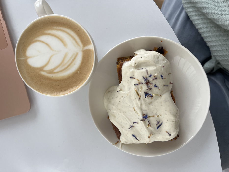 An overhead shot captures a coffee and cake arrangement on a white table in Callbach, Germany. A white ceramic mug filled with a latte featuring a detailed rosetta latte art pattern is positioned in the upper right. Below and to the left of the mug, a white ceramic plate holds a slice of cake, possibly carrot cake, topped with a substantial layer of white, speckled frosting. This frosting is garnished with scattered dried flower petals, primarily blue and light brown/orange hues. In the upper left corner, a portion of a rose-gold colored laptop is visible. The lower right section of the frame shows a light blue fabric item, likely clothing or a blanket. No individuals are visible.