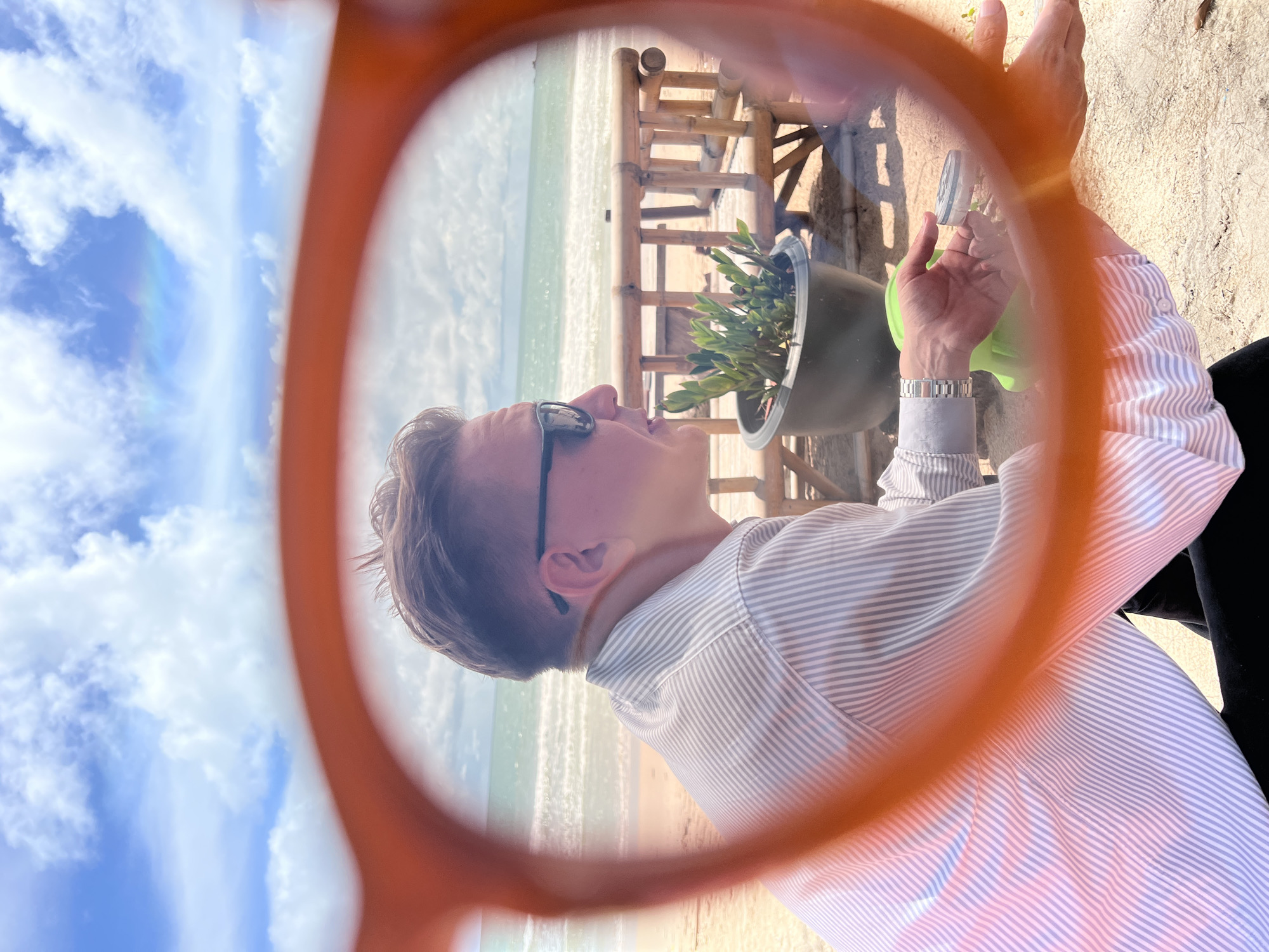 A person is sitting on a sandy beach in front of a body of water and a bamboo structure. They are wearing a striped button-down shirt and sunglasses and appear to be holding a cigarette and a small round container. The foreground is blurred by an orange lens, possibly from glasses. The sky is blue with white clouds, and there is a faint rainbow visible.