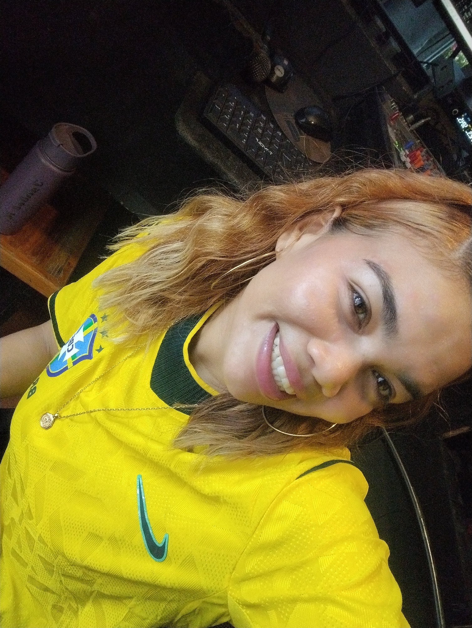 A close-up, low-angle selfie shows a young woman with blonde-red hair and brown eyes smiling at the camera. She is wearing a bright yellow soccer jersey with a green trim and a crest on the left side of the chest. A thin gold chain with a circular pendant rests on the jersey. The background is dark and out of focus, suggesting an indoor setting. To the left, a dark purple cylindrical object, possibly a water bottle, is visible. Behind her, a keyboard and a computer monitor are partially visible. The overall impression is that of a casual self-portrait, likely taken in a home or office environment, with no specific identifiable landmarks or features that would place it in Porlamar, Venezuela.