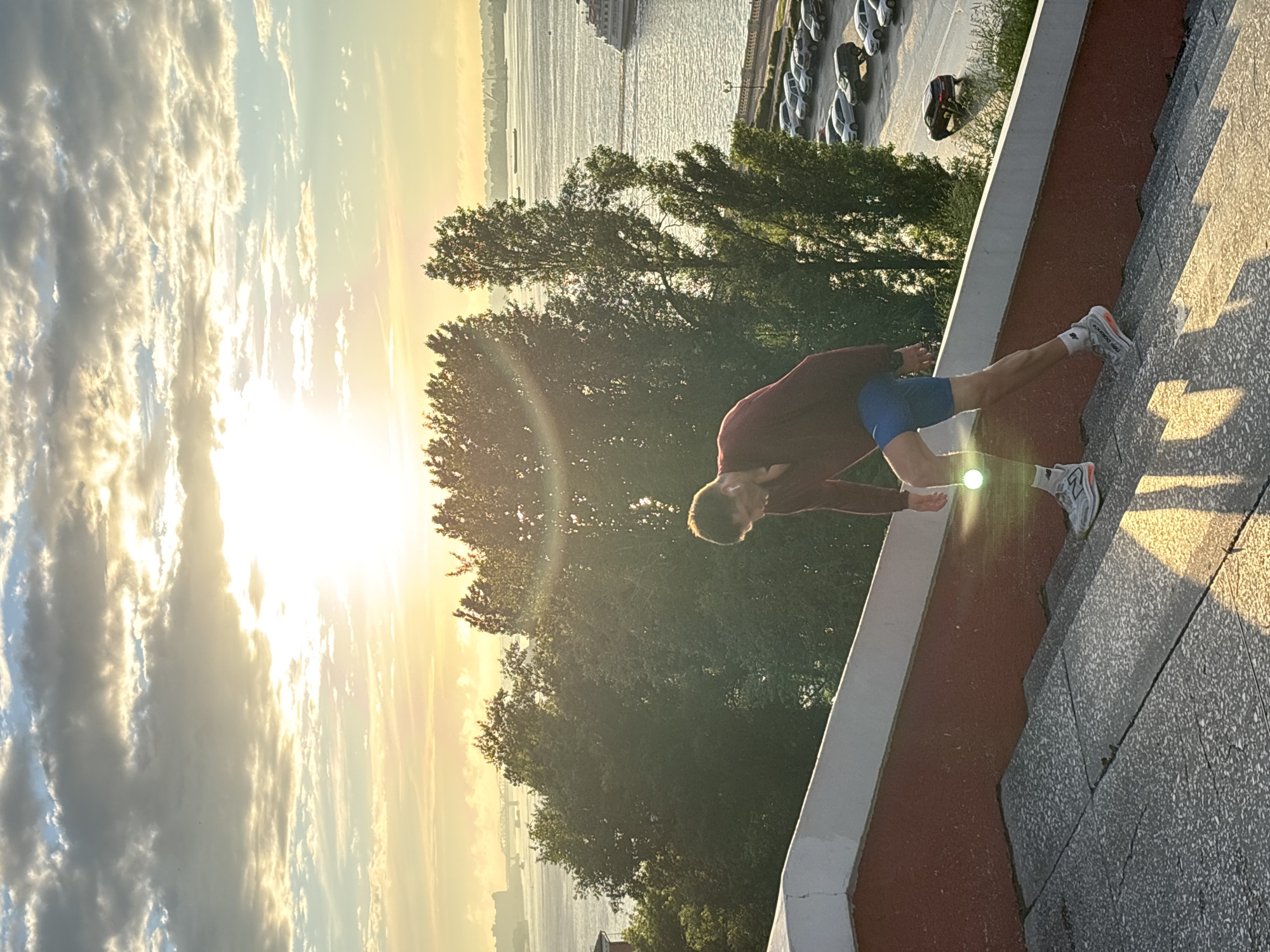A man in athletic wear is ascending a wide, outdoor staircase during daylight. He is wearing a maroon long-sleeved shirt, blue shorts, and grey athletic shoes. His body is angled forward, and his right leg is extended upwards on a step. Sunlight glints off his shoe and casts long shadows across the staircase. The staircase has a red-painted wall to its left and is made of large, grey, speckled concrete slabs.
In the background, lush green trees are abundant. Beyond the trees, a wide body of water, likely the Volga River, is visible, with a boat or ship sailing on it. Further in the distance, a bridge spans the river, and the silhouette of buildings can be seen on the horizon. A parking lot filled with cars is situated along the embankment. The sky is partly cloudy, with the sun shining brightly through the clouds, creating a luminous effect. The scene appears to be set in Nizhny Novgorod, Russia, based on the distinctive geographical features and architectural elements.