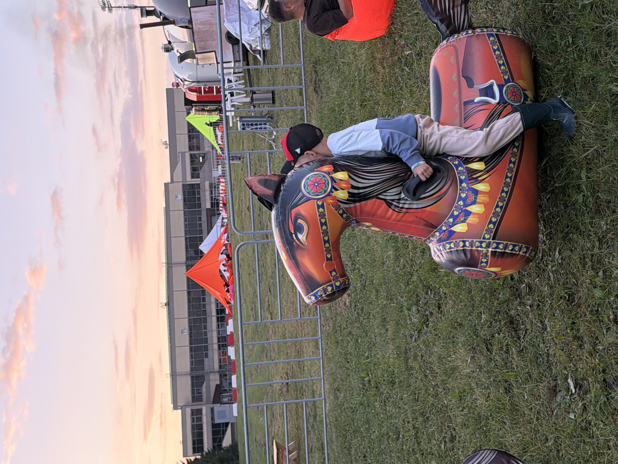 An outdoor scene at dusk, likely in a park or open field, with a soft pink and orange sky. In the foreground, a young person is playing on a large, inflatable brown and red horse. The child is wearing a light blue and gray long-sleeved shirt, light beige pants, and dark green boots, with a black baseball cap. The child's face is partially obscured, turned towards the horse's neck. The inflatable horse is decorated with traditional-looking patterns. In the midground, a metal fence separates the grassy area from a paved path and a large building in the background. Several colorful tents, including a bright orange one, are visible near the building. To the right, a person is sitting on a bright orange beanbag chair. The overall atmosphere suggests a relaxed, possibly festive, event.