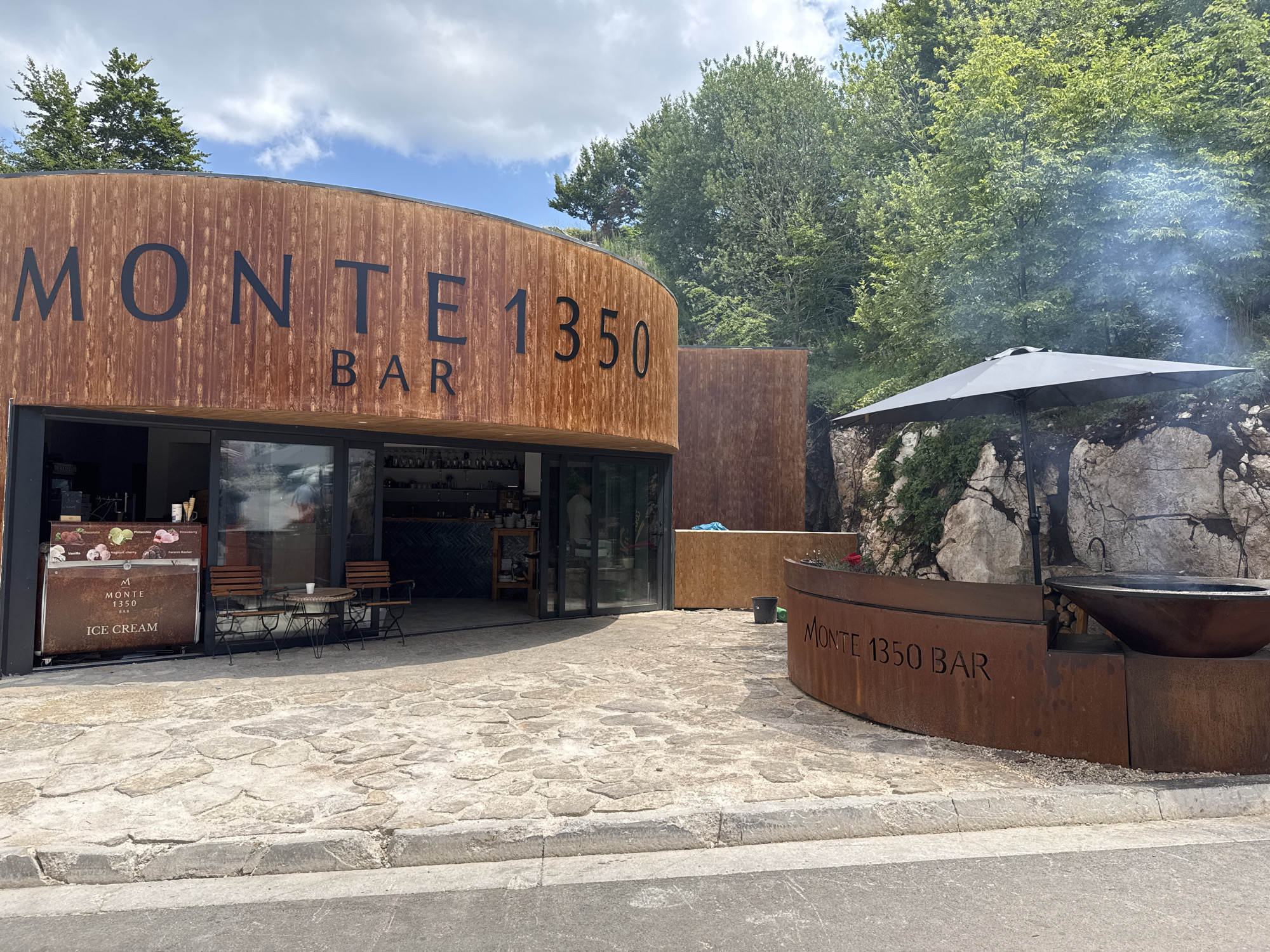 The image shows a modern, rustic-style bar called "MONTE 1350 BAR" located in Žanjev Do, Montenegro. The building's facade is constructed from weathered wood, with large black lettering spelling out the bar's name. To the left of the entrance, an ice cream display is visible. Seating for patrons, consisting of two chairs and a table, is placed on a cobblestone patio in front of the establishment.
To the right of the building, a large, curved metal structure, also bearing the "MONTE 1350 BAR" inscription, is present. This structure appears to house a large, dark cauldron or cooking vessel, with smoke rising from it, suggesting a barbecue or outdoor cooking activity. An umbrella provides shade over this area. The setting is nestled against a rocky hillside, with lush green trees and foliage forming a backdrop. The sky is partly cloudy, indicating daytime. The overall impression is one of a contemporary outdoor establishment designed to blend with its natural surroundings, offering a place for relaxation and refreshments.