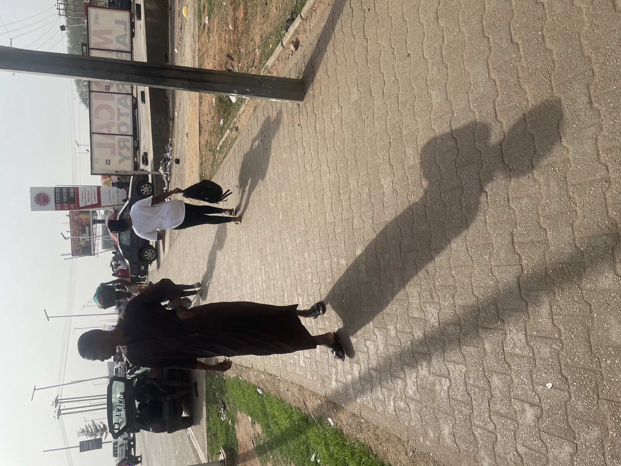This image, if viewed rotated 90 degrees clockwise, depicts a bright, sunny street scene, likely during mid-morning or late afternoon, as indicated by the long shadows cast across the ground. The setting is an urban or semi-urban area, consistent with Abuja, Nigeria.
On a sidewalk paved with interlocking concrete pavers, two individuals are walking. One person, seen from behind in a white t-shirt, dark leggings, and sandals, carries a black backpack. Their elongated shadow stretches across the pavement to the right. Ahead of them, walking towards the camera, is another person dressed in a long, dark robe-like garment and sandals, also casting a prominent shadow.
To the left of the sidewalk is a road with several vehicles. A dark car is partially visible, and further back, a large truck with a white container on its bed can be seen. The container displays mirrored text, which, when reversed, appears to read "MAJOR FACTORY."
A tall, vertical sign structure is prominent on the left side of the frame. It features the NIPCO logo at the top and lists fuel prices: PMS 1292:00, AGO 1950:00, and LPG 00:00. Below these prices, services such as "Laube Bay," "Mini Mart," and "Lubricant" are advertised. Another smaller sign in the background partially shows the word "STANDARD."
The scene captures typical daily activity, with people walking and vehicles on the road. Patches of dry grass and some scattered litter are visible alongside the sidewalk and road, indicating a bustling street environment under a clear sky.