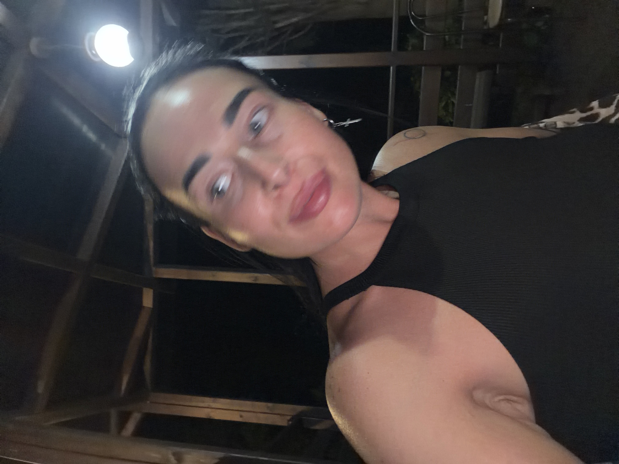 A woman is captured in a close-up, selfie-style shot at night, appearing to be outdoors under a wooden structure. She is looking to her left, with her mouth slightly open. She has dark hair pulled back, prominent eyebrows, and is wearing a black sleeveless top. A silver cross earring is visible in her left ear. A circular tattoo is partially visible on her upper right arm. The background is dark, with some wooden beams and a lit bulb overhead suggesting a covered outdoor area, possibly a gazebo or patio. The overall impression is that she is in a casual, nighttime setting, possibly in the city of Sochi, Russia.