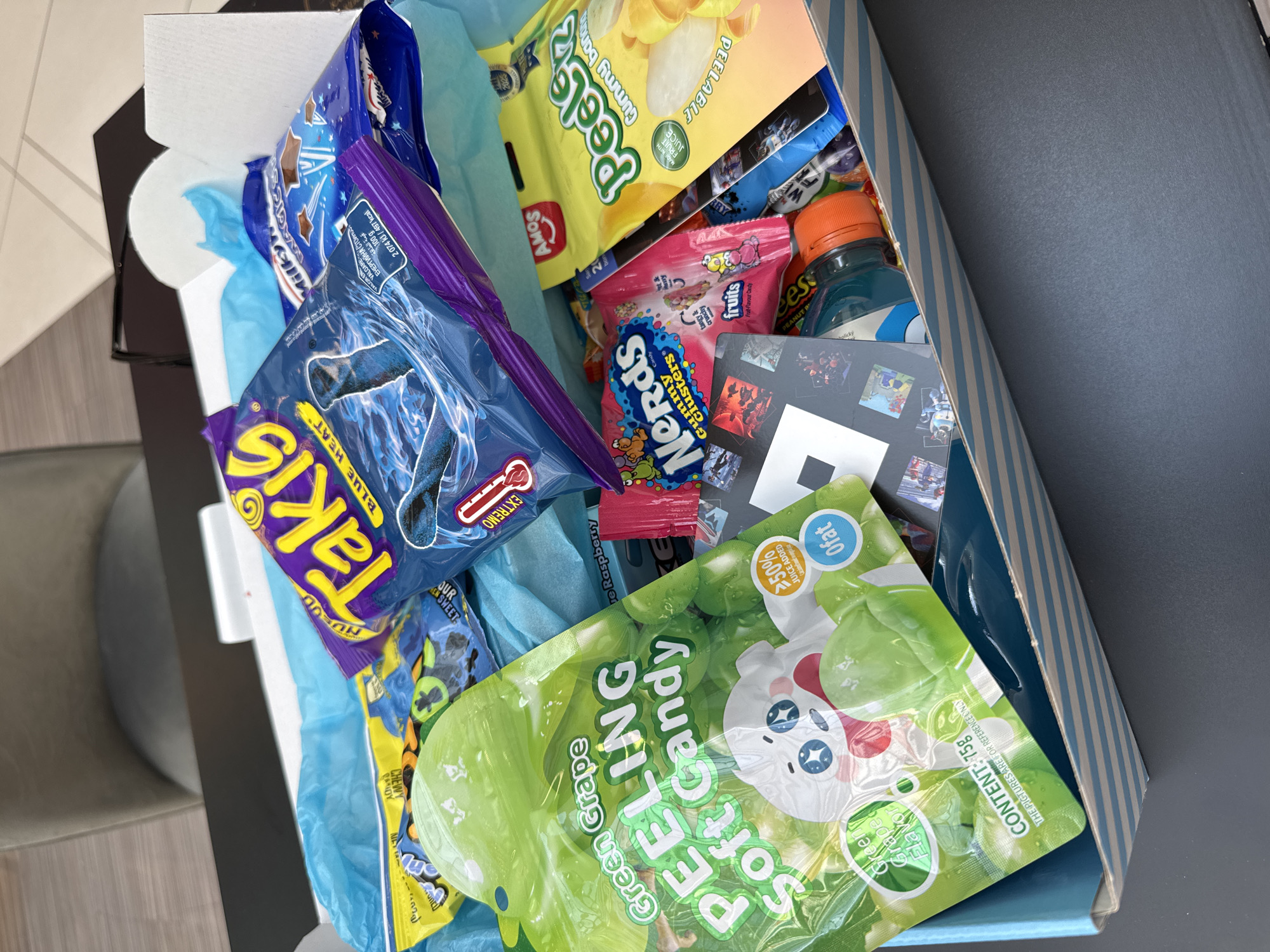 An overhead, slightly angled photograph captures an open rectangular cardboard box filled with various confectionery items and snacks, positioned on a dark gray table. The interior of the box is lined with crumpled light blue tissue paper.
Dominating the contents are a large blue bag of "Takis Blue Heat" rolled tortilla chips, visible centrally, and a prominent green pouch of "Green Grape PEELING Soft Candy," featuring a white cartoon ghost and marked with "CONTENT: 75g" and "0fat." Also clearly visible are a yellow pouch of "AMOS Peelerz Gummy Banana" with the text "PEELABLE" and "MADE WITH FRUIT JUICE," a pink bag of "Nerds Gummy Clusters," and a partially obscured blue bag of "Milky Way Magic Stars." Additional items include an unidentified small bottle with an orange cap and several small, rectangular stickers depicting various images, arranged near the bottom of the box.
The background, out of focus, reveals a light-colored chair and a light-toned floor. No individuals are present in the image. There are no identifiable visual cues or unique markers within the photograph that indicate a specific geographic location such as Riga, Latvia.