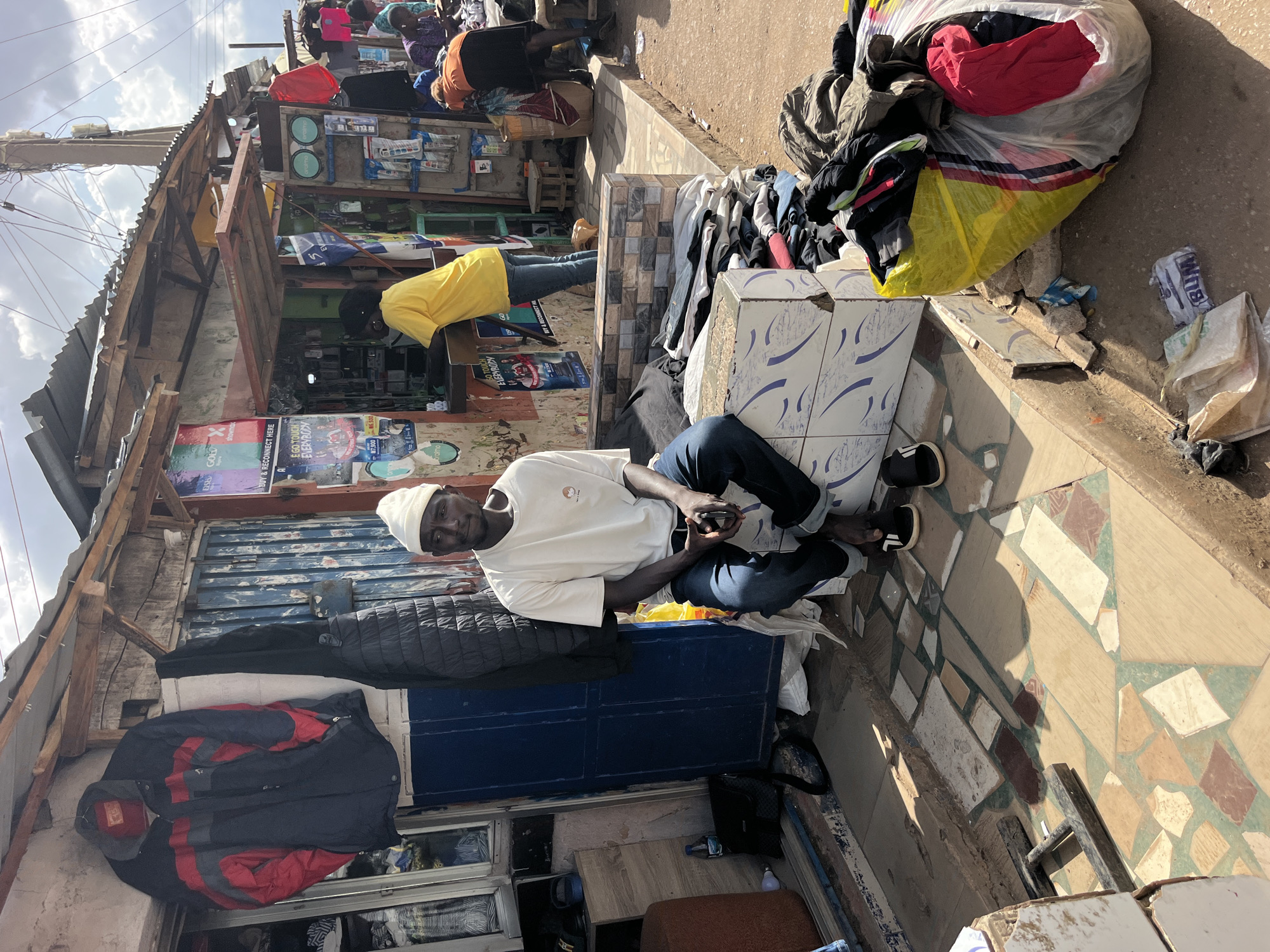 In an outdoor market scene, a Black man wearing a white t-shirt and blue jeans, and a white head covering, sits on a low wall. He is looking towards the camera. Behind him, various stalls display clothing and goods. To his left, a stall features a blue door and jackets are hanging. In the background, there are more stalls, some covered with corrugated metal, and people can be seen browsing or working. Large bags of clothing are piled up to the right of the man. The ground is tiled with a mosaic of colored stones. Sunlight suggests it is daytime. The scene appears to be a bustling marketplace. Visible text includes "ESH GOL", "BUY & RECONNECT HERE", "E GO TOUCH EVERYBODY", "79006,500", and "BUM".