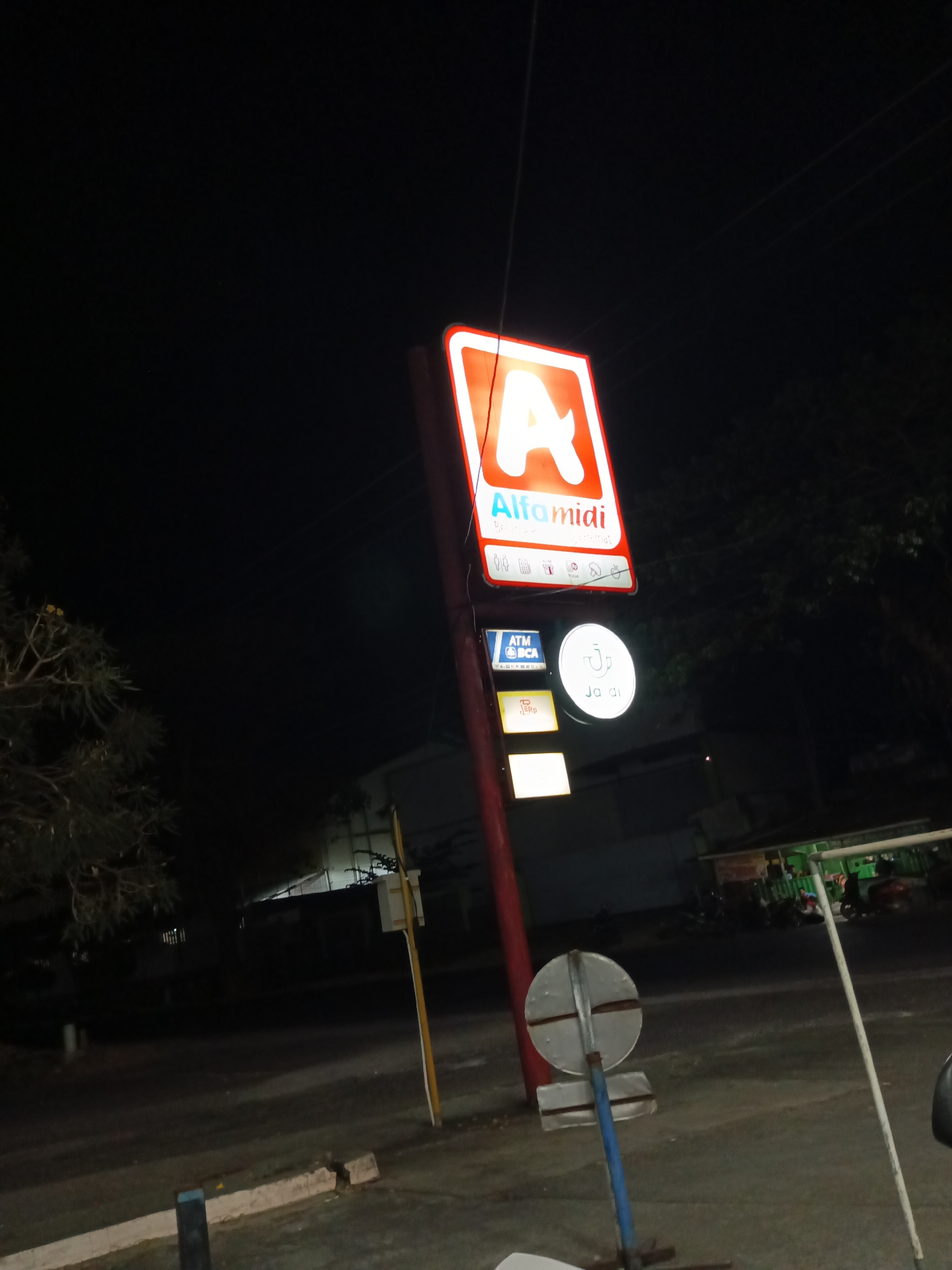A lit, red and white sign for "Alfamidi" is visible at night, positioned on a tall red pole. Below the main sign, there are several smaller illuminated signs, including one for "ATM BCA", a circular sign with a cup graphic and "Ja di" written on it, and a yellow rectangular sign labeled "eRp". To the left of the pole, a tree with sparse foliage is present. The background shows a dark, unlit area with some structures and trees. A circular, reflective traffic sign is also in view, mounted on a blue and silver pole. The ground appears to be asphalt or pavement.