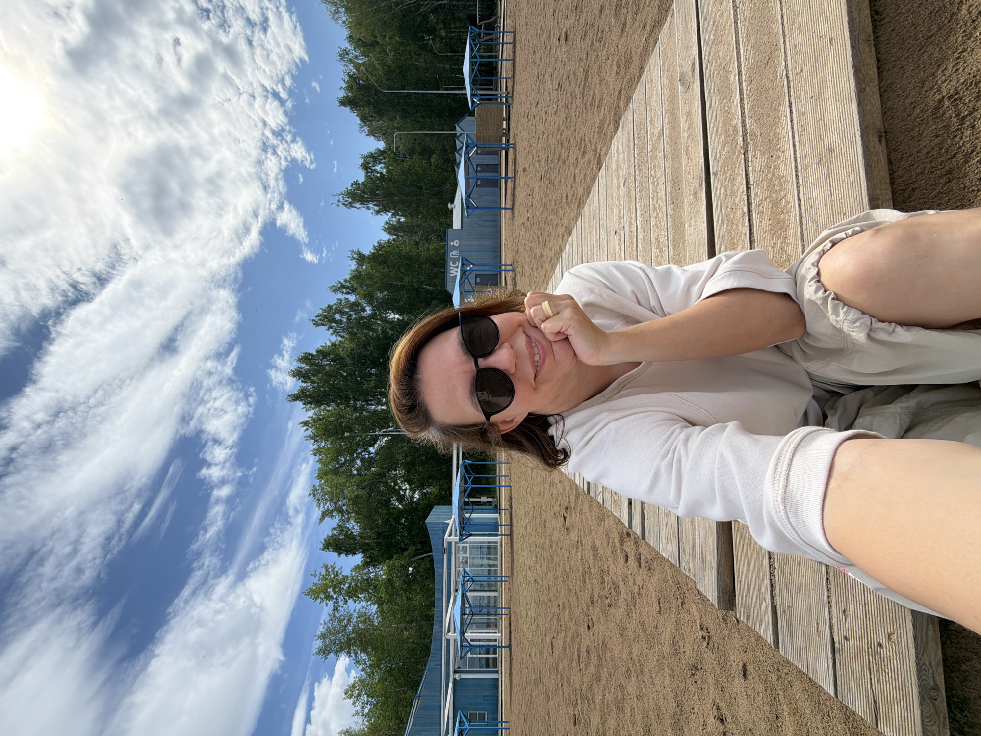 A woman wearing sunglasses and a light-colored long-sleeved shirt is seated on a wooden boardwalk on a sandy beach. She is smiling and resting her chin on her hand. In the background, there are trees, a light blue building with tents, and a cloudy sky. This appears to be the beach area in the city of Chaikovsky, Russia.