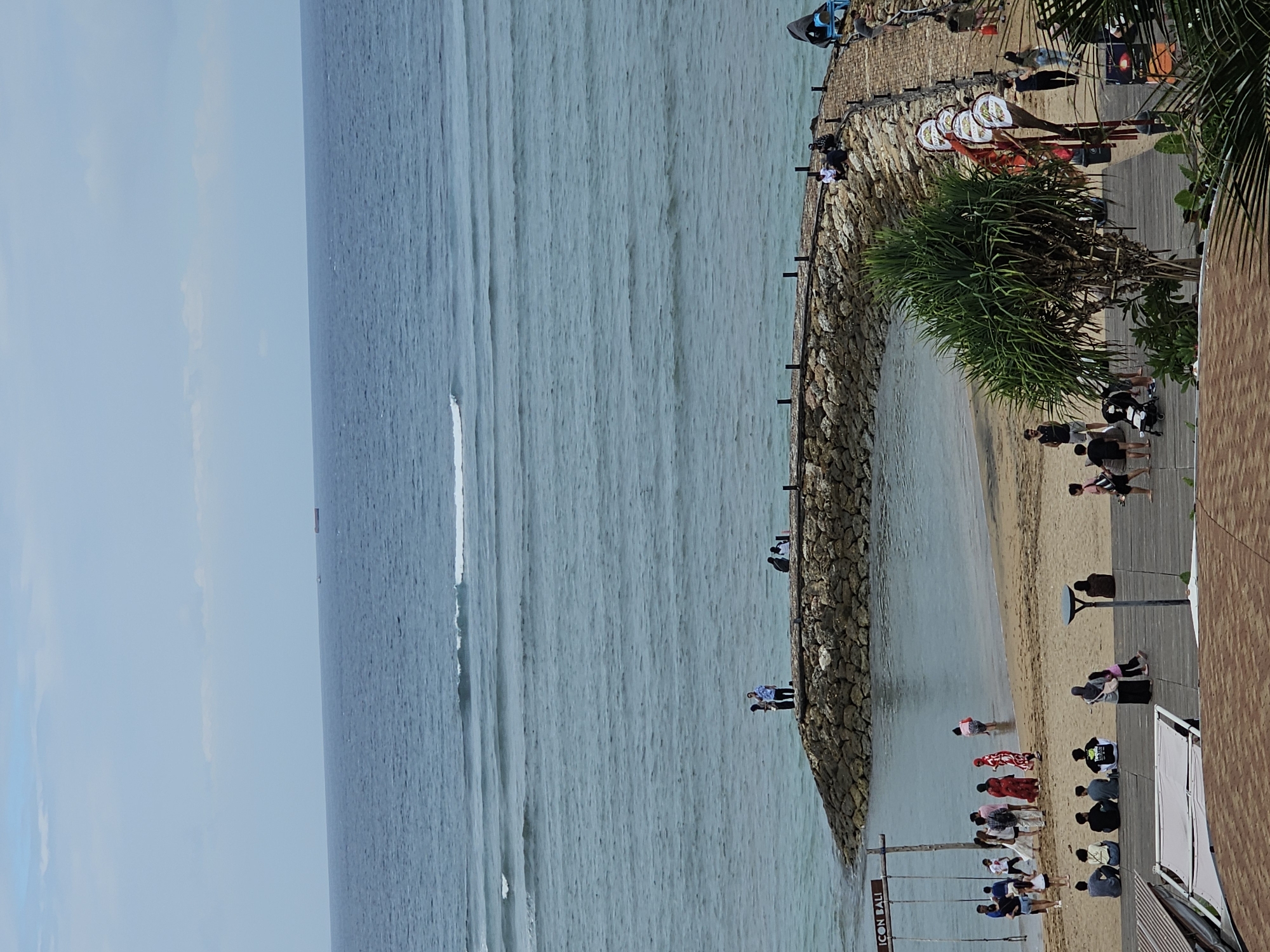 The image depicts a coastal scene at Seminyak Beach in Denpasar, Indonesia. The turquoise ocean stretches to the horizon under a partly cloudy sky. Gentle waves roll towards a sandy beach where numerous people are visible.
In the foreground, a wooden boardwalk runs parallel to the beach, with several individuals sitting and standing on it. To the left, a large wooden swing set with the text "ICON BALI" is prominent. A group of people are gathered near the swing set, some standing and some interacting.
Further down the beach, near the water's edge, more people are present. Some are wading in the shallow water, while others are standing or walking along the sand. A curved, stone jetty extends into the ocean from the right side of the frame, with a few individuals standing and sitting on it. On the right side of the image, decorative lanterns hang from poles along the boardwalk. A lush green pandanus tree stands near the boardwalk, adding to the tropical ambiance. In the far distance, a large ship is visible on the ocean. The scene conveys a relaxed, leisurely atmosphere typical of a beach resort.