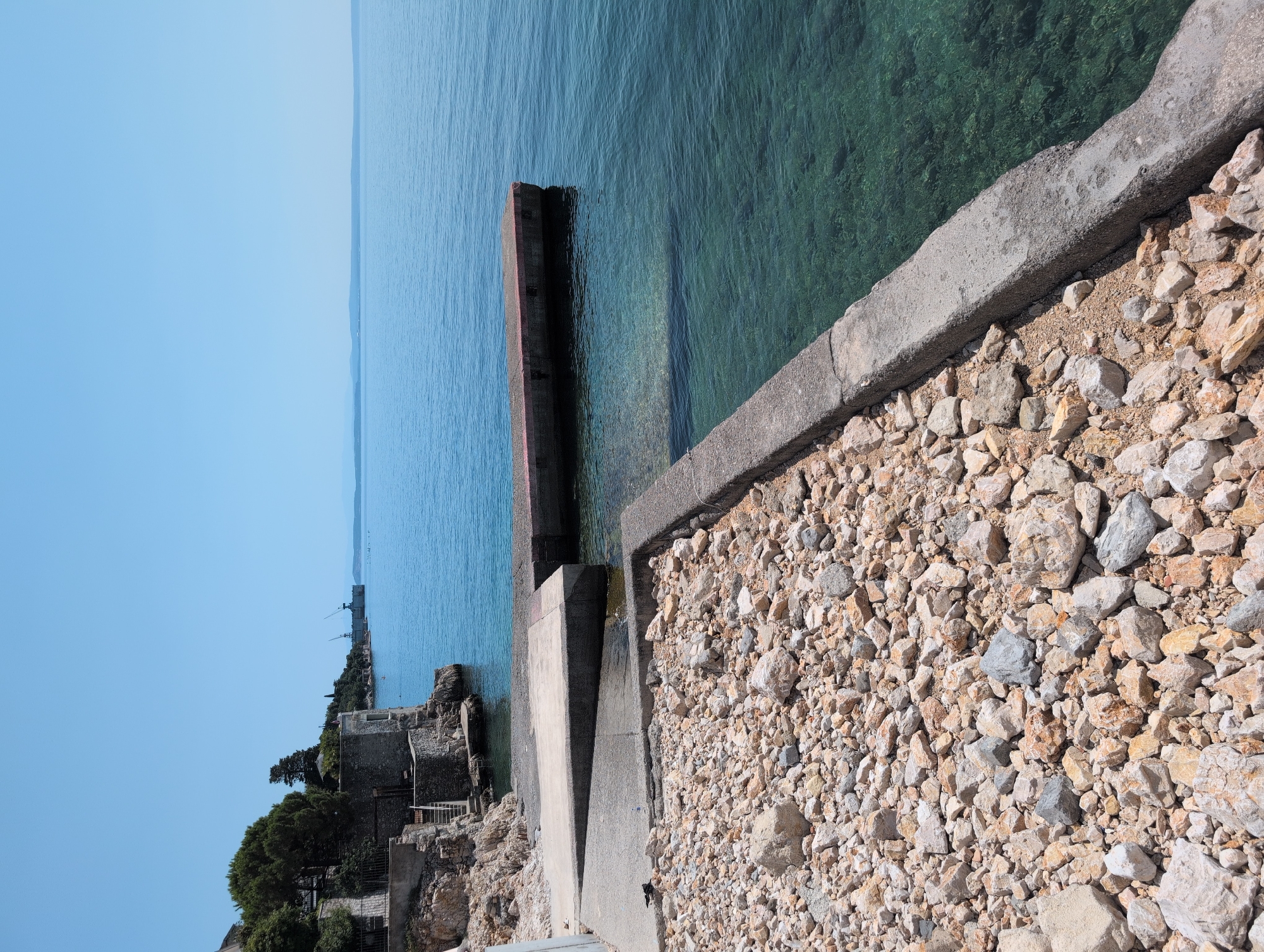 The image depicts a coastal scene in the city of Rijeka, Croatia, likely near the industrial port area given the presence of cranes in the background. In the foreground, a concrete pier juts out into the clear, blue-green water. The pier is surrounded by a rocky shoreline. To the left, a stone building and stairs are visible, suggesting a historical or residential structure. The water is calm and transparent, allowing a view of the seabed. In the distance, industrial cranes and structures are faintly visible against the horizon, indicating the proximity of a port. The sky is clear and blue, suggesting a sunny day.