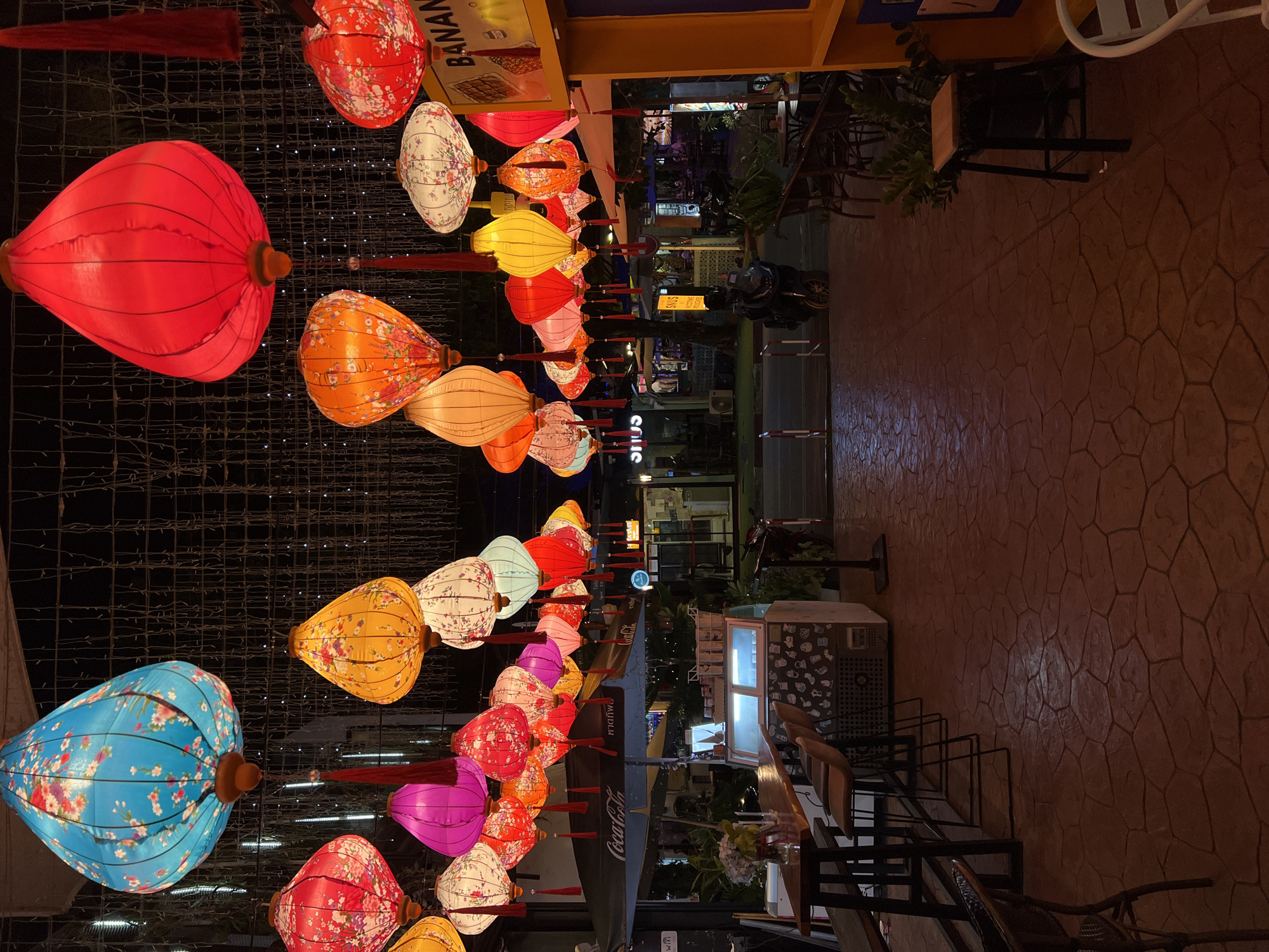 The image depicts a street scene at night, adorned with numerous colorful, floral-patterned paper lanterns hanging overhead. The lanterns, in shades of blue, red, pink, yellow, and white, are illuminated and create a festive atmosphere. Below the lanterns, a pathway paved with reddish-brown hexagonal stones leads towards the background. On the left side of the pathway, there is an outdoor seating area with wooden tables and chairs, a counter, and an ice cream display. A Coca-Cola umbrella is visible above the seating area. On the right side, there are more tables and chairs, some foliage, and a yellow structure with signage that includes the word "BANANA". In the mid-ground, a street with a few parked vehicles and building storefronts is visible. The overall scene suggests a lively, possibly commercial or entertainment district in a city at night.