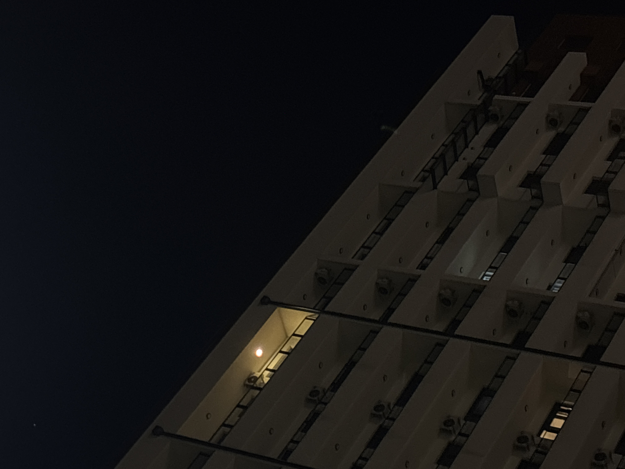 The image depicts a section of a building at night. The structure is modern, featuring a repeating pattern of recessed rectangular windows or spaces. The facade appears to be made of light-colored concrete or stone. The lighting is dim, with one window illuminated by an internal light source. A faint light in the sky, likely a star or planet, is visible in the upper left corner. Details such as ladders and possibly air conditioning units can be seen on the building's exterior. The location is likely Belgrade, Serbia, as the building closely resembles the Genex Tower, a prominent landmark in the city.