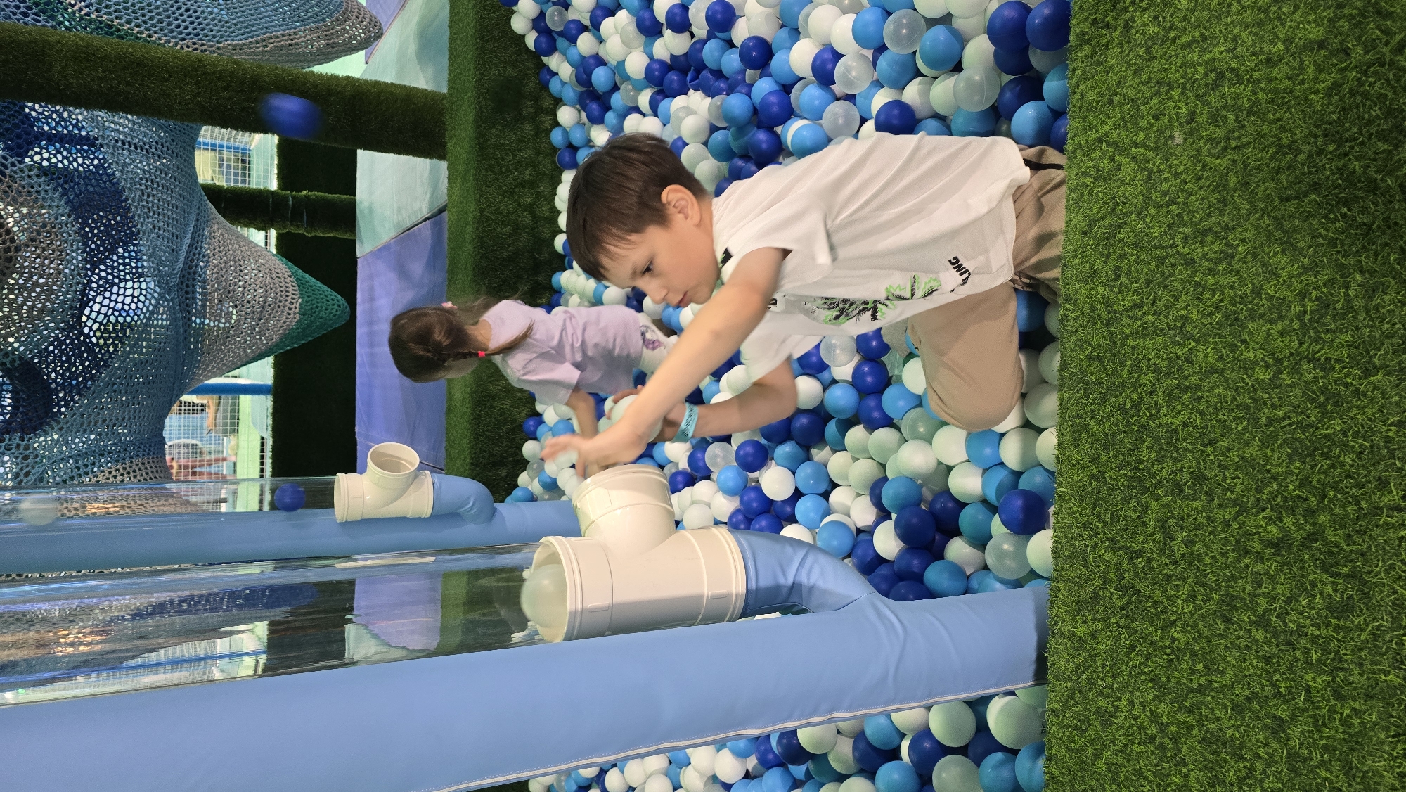 This image captures an indoor children's play area located in Izhevsk, Russia. The scene is dominated by a large ball pit filled with plastic balls in shades of dark blue, light blue, and white.
In the foreground and central-left, a prominent light blue padded vertical structure stands next to a clear plastic tube assembly. This tube system incorporates white PVC pipe fittings, including elbows and T-junctions, designed for moving balls, with several balls visible within its transparent sections.
A young boy, approximately 5-8 years old, with short light brown hair, is positioned on the right side of the image, kneeling or squatting in the ball pit. He is wearing a white short-sleeved t-shirt with a subtle graphic design and light brown or khaki shorts. He has a blue wristband on his left arm. The boy is actively engaged with the ball-moving tube system, holding a white ball with both hands as if in the process of placing it into or retrieving it from one of the white pipe openings. His gaze is directed towards the ball and the mechanism.
Slightly behind the boy and to the left, a younger girl, approximately 3-5 years old, with light brown hair styled in pigtails, is partially visible within the ball pit. She is wearing a light purple t-shirt and patterned shorts, facing away from the camera towards the background.
The floor in the immediate foreground consists of vibrant green artificial turf, separating the viewer's perspective from the ball pit. In the background, additional play structures are visible, including expansive blue and grey mesh netting, characteristic of climbing or contained play zones. A vertical support column or structure covered in green artificial grass also rises in the background on the right, blending with the netting. The overall environment suggests an artificial, brightly lit indoor setting.