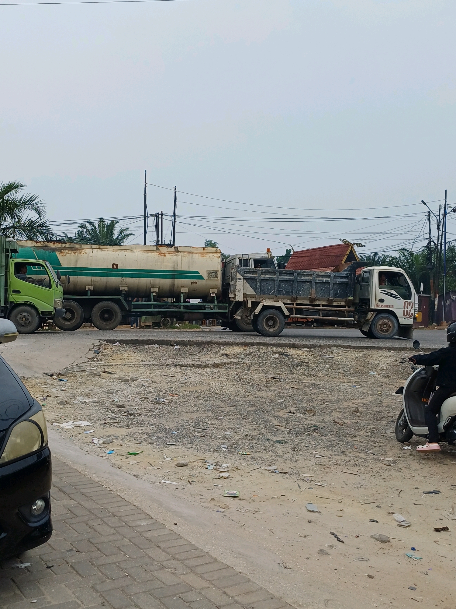 The image depicts a roadside scene in Dumai City, Indonesia, on a cloudy day. Two trucks are the primary subjects, positioned on a paved road. The truck on the left is a green tanker truck with a white and green horizontally striped tank. A driver is visible in the cab. To the right of the tanker truck is a white flatbed truck with a dark grey cargo bed. This truck has the number "02" prominently displayed on its side in red. A person is visible in the driver's seat of the flatbed truck. The immediate foreground shows a black car partially visible on the left, and a white scooter with a rider dressed in black and pink shoes on the right. The ground beside the road is unpaved and appears to be covered in gravel and debris. In the background, palm trees and power lines are visible, along with a building with a distinct red, pointed roof. The overall impression is one of an urban or semi-urban environment with ongoing traffic.