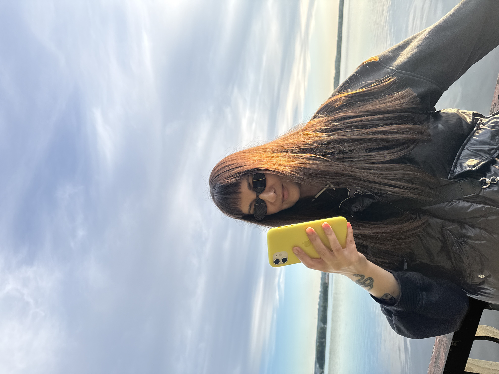 A woman stands outside in daylight holding a yellow mobile phone. She has long brown hair with bangs and wears sunglasses and a black jacket. A body of water stretches behind her, with land visible in the distance. The sky is cloudy. Due to lack of data, it is impossible to definitively identify the location as Votkinsk, Russia.