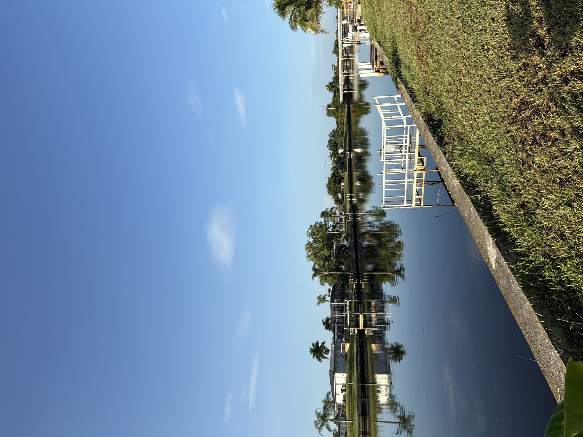 The image shows a calm canal with reflections of houses, palm trees, and a bridge. A white boat dock extends into the water on the right side of the canal, and another structure is visible further down the canal. The sky is clear blue with a few wispy clouds. The banks of the canal are lined with green grass and some tropical foliage. The scene appears to be a residential area in a warm climate, possibly Florida, given the presence of palm trees and canals.