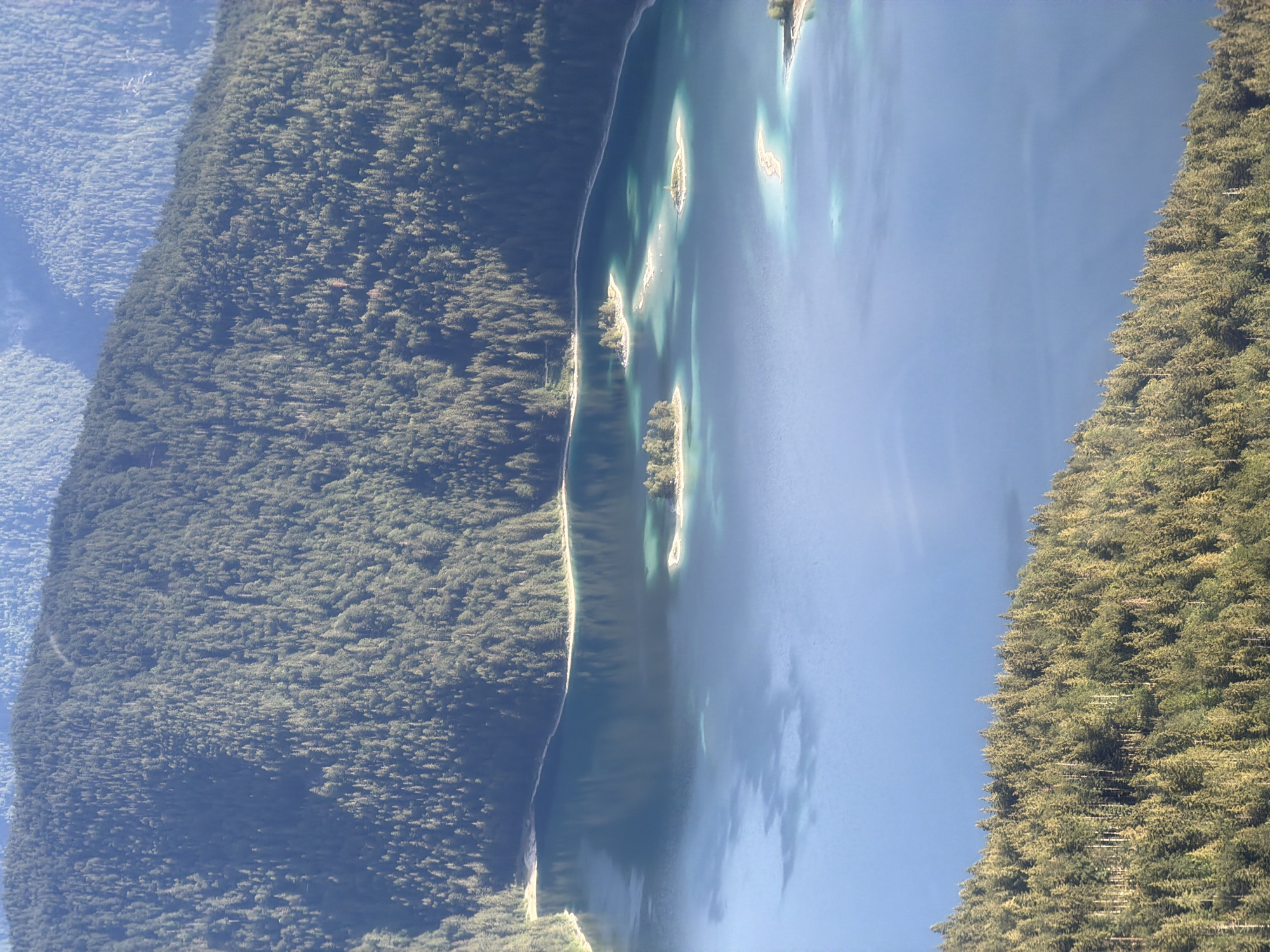 This is an aerial, eye-level view of the Eibsee lake in Grainau, Germany. The lake is a deep blue color and has several small, tree-covered islands scattered across its surface. The surrounding landscape is dominated by dense evergreen forests that cover the steep mountain slopes. The sun is shining, casting shadows and highlighting the textures of the trees and water. The overall scene is one of natural beauty and tranquility.