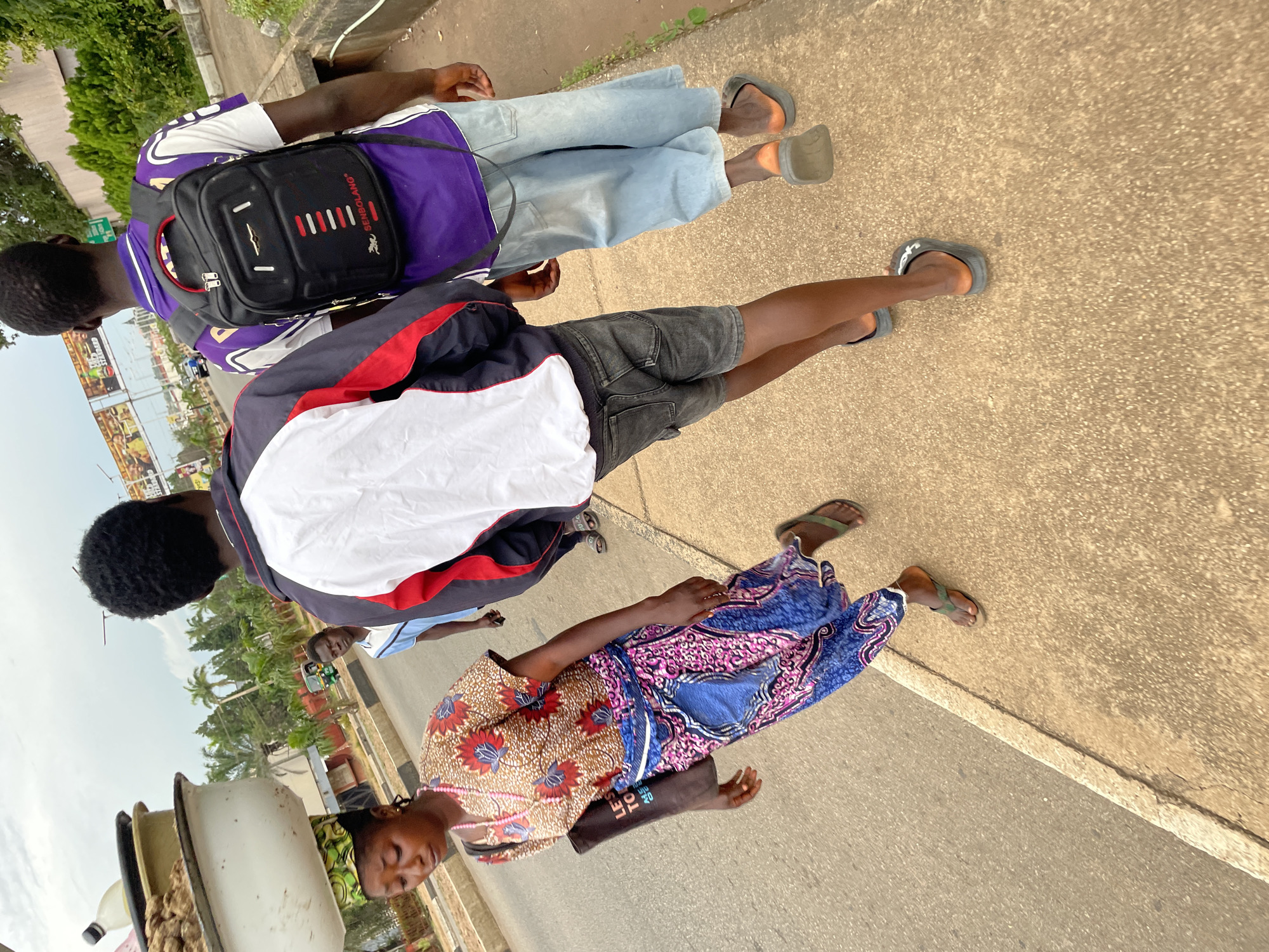 The image depicts a street scene in Jos, Nigeria. In the foreground, a woman is walking on a paved sidewalk, carrying a stack of containers on her head. She is wearing a colorful patterned dress and a pink beaded necklace. Next to her, two young men are walking in the same direction. The man on the left is wearing a dark jacket with red and white trim over a white shirt and dark shorts. The man on the right is wearing a purple and white shirt with a black backpack and light-wash baggy jeans. They are walking on the sidewalk adjacent to a road with vehicles visible in the background. Palm trees and vegetation line the side of the road, and billboards are visible in the distance. The sky is overcast.