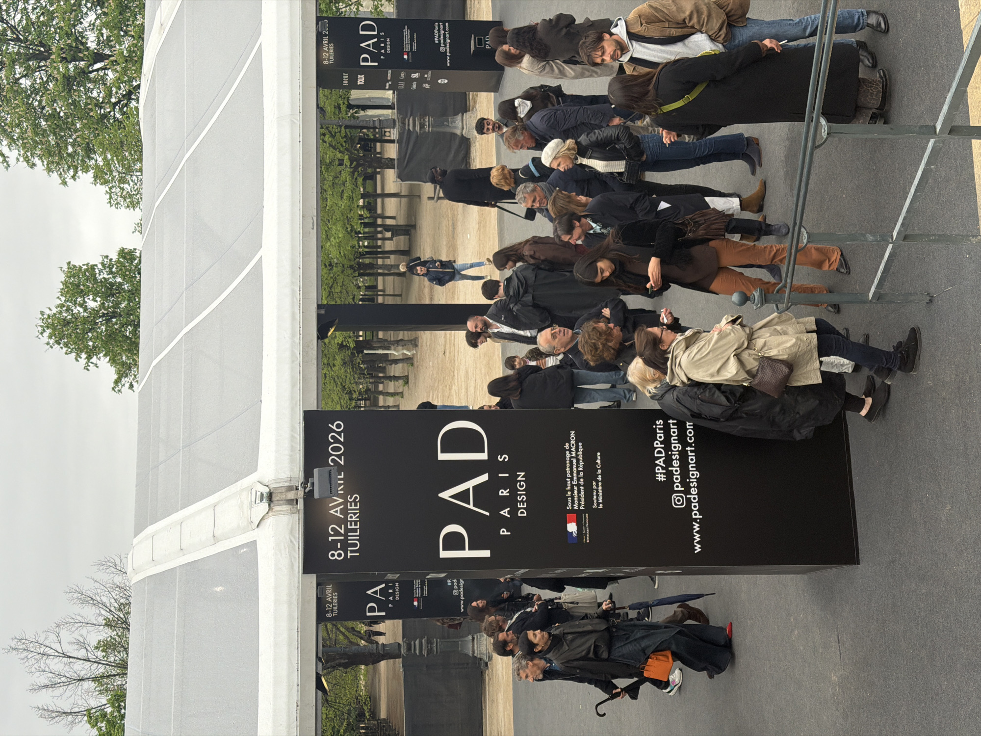 Here is a description of the image:
The image shows a crowd of people queued outside a black sign for the "PAD Paris Design" event, which is taking place from April 8-12, 2026, in the Tuileries Garden, Paris, France. The sign also bears the logos of various sponsors, the social media handles and website for the event. The people are standing on a grey surface and are dressed in a variety of casual clothing. A partially visible tent structure can be seen in the background. There are trees adjacent to the tent.