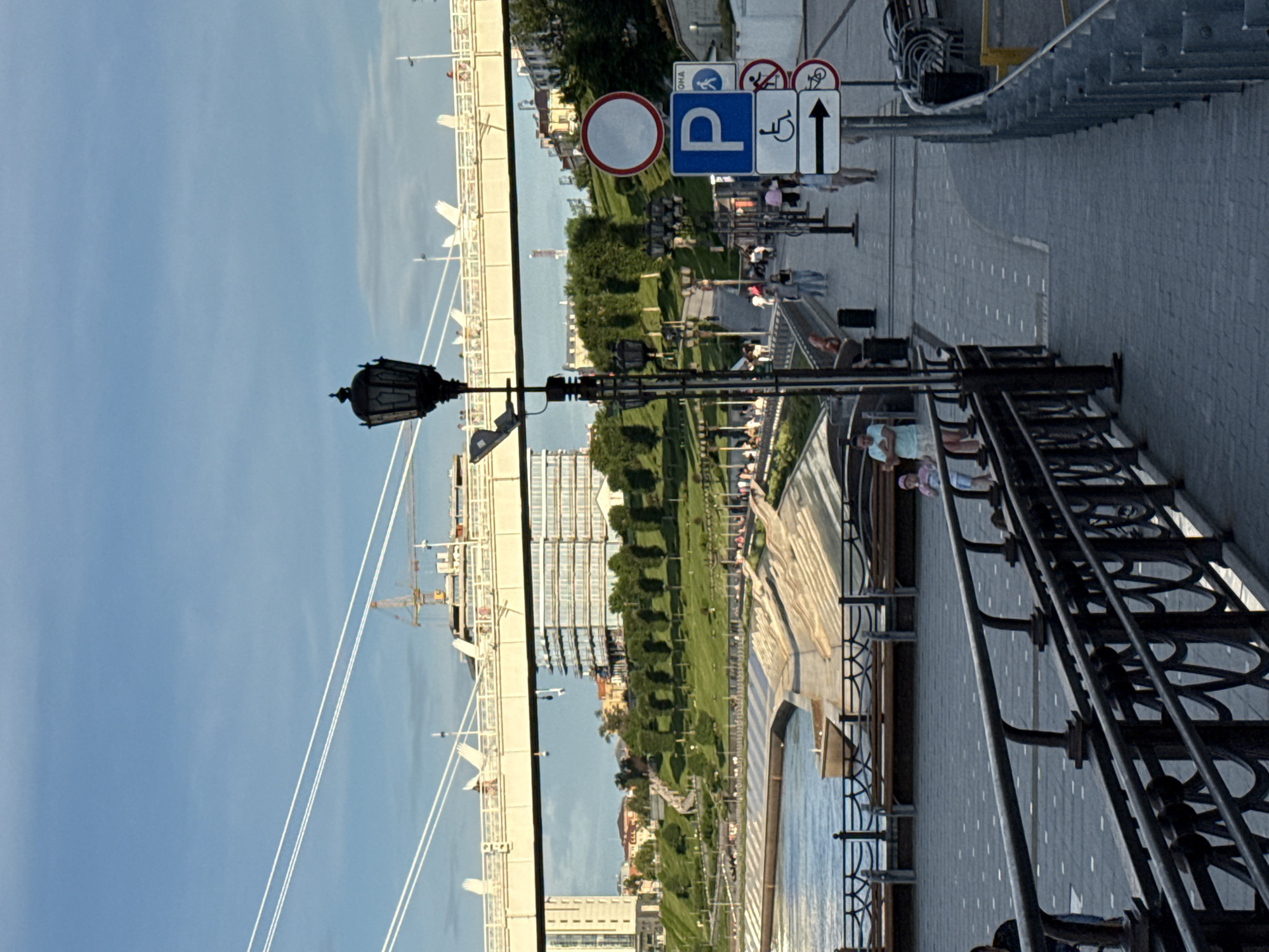 The image depicts a view of the Tyumen Embankment in Tyumen, Russia. The foreground features a dark metal railing along a paved pedestrian walkway. To the right, a series of traffic signs are visible, including a blue "P" for parking, an accessibility symbol, and a directional arrow pointing right, indicating a path for bicycles. Further back, people are walking along the embankment. Beyond the walkway, a tiered structure with benches overlooks a body of water. In the mid-ground, a grassy slope is lined with trees and a winding path, leading up towards buildings. A large modern building with many windows dominates the background, with a bridge extending across the frame above it. Construction cranes are visible behind the modern building, suggesting ongoing development. The sky is a clear blue with some wispy clouds. The overall scene suggests a public recreational area on a sunny day.