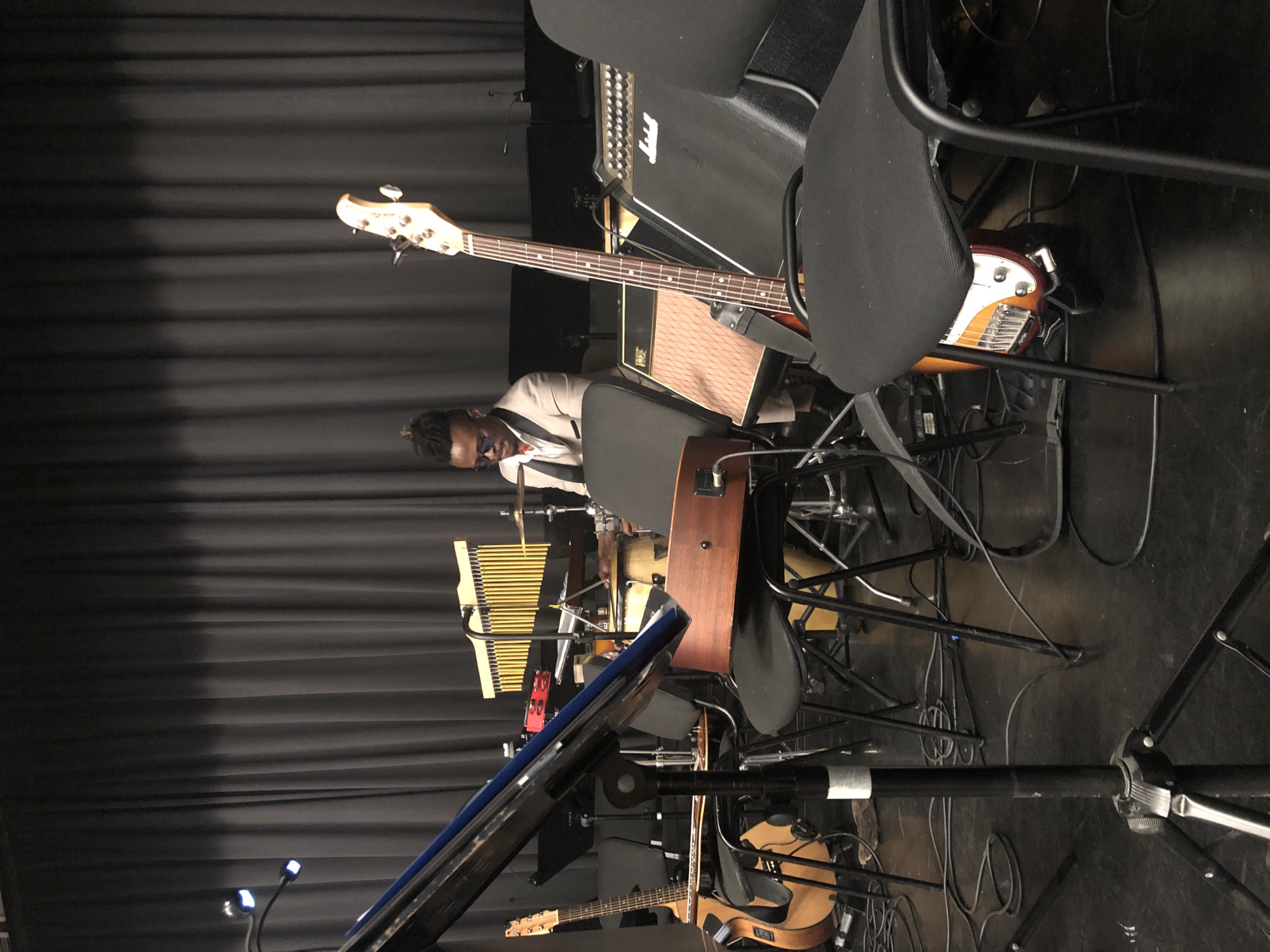 A musician, appearing to be a Black male with dreadlocks and wearing a light-colored suit with sunglasses, is seated behind a drum set and other musical instruments, including a xylophone, in front of a dark, pleated curtain. In the foreground, a bass guitar stands upright, its neck extending towards the top of the frame. Several chairs are positioned around the stage, some with musical instruments visible beneath them. Various cables snake across the floor, connecting the equipment. The lighting appears to be stage lighting, with two visible spotlights attached to a stand in the lower left corner. The overall setting suggests a performance or rehearsal space. There is no indication of the location being Moscow, Russia.