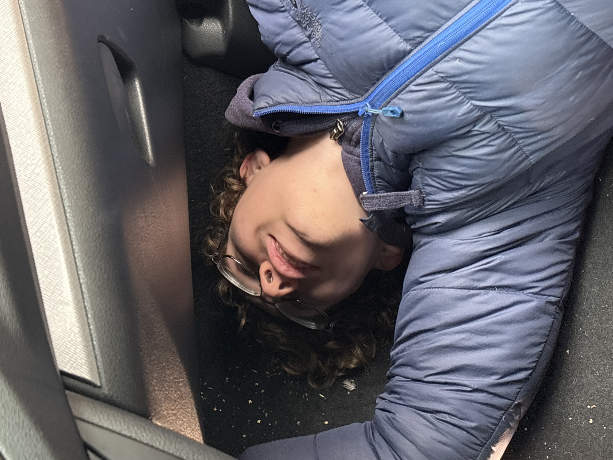 A young person, wearing glasses and a blue puffy jacket, lies on a dark, possibly carpeted, surface next to a gray, textured wall panel, possibly inside a vehicle. There is no information to suggest the location is Perm, Russia.