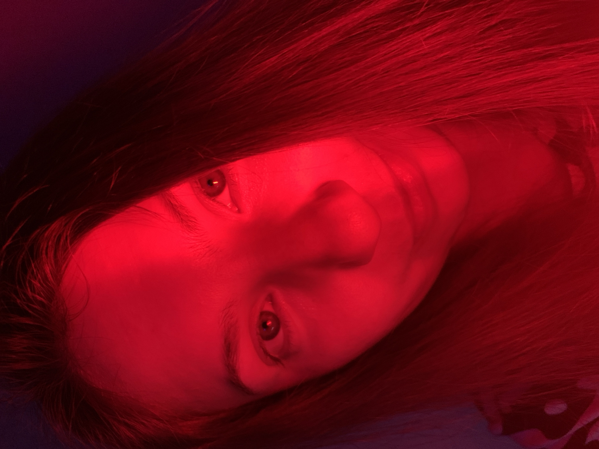 The image is a close-up, high-angle shot of a person's face illuminated by red light, with their hair falling over the right side of their face. The person's left eye is clearly visible, while the right side of their face is partially obscured by shadow and hair. The red lighting creates a strong contrast and casts dramatic shadows, highlighting the contours of the face. The background is dark and indistinct. The image does not contain any specific elements that would allow for identification of its location within Chaikovsky, Russia.