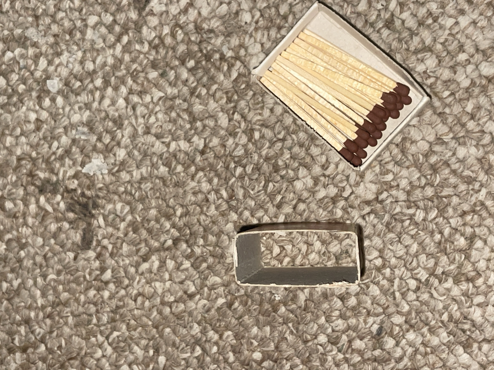 The image shows a close-up of a textured, beige carpet. On the carpet, there are two objects: a small, empty, rectangular cardboard matchbox, and a single, detached metal frame, possibly part of a picture frame or a decorative item, that is hollow and rectangular in shape. The matchbox is filled with several wooden matches with brown tips, laid out in a fan-like arrangement. The scene does not depict any specific location in Jalingo, Nigeria, nor does it include any people, actions, or interactions.
