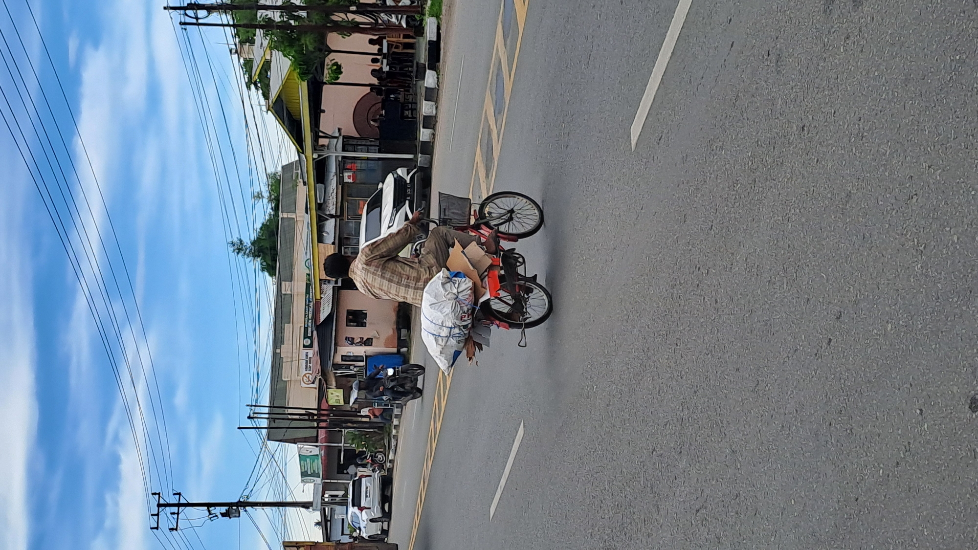 A man rides a red bicycle laden with goods down a paved road in Julikeude Dua, Indonesia. The man, wearing a plaid shirt and dark pants, is positioned in the center of the frame. The bicycle's rear rack is stacked high with a large, white, tarp-wrapped bundle and several pieces of cardboard. A black basket is attached to the handlebars. The road has yellow and white lane markings. In the background, several businesses with awnings and signage line the street, including a building with the text "SIRIBUEH MEDICA". Several other vehicles, including a white SUV and a motorcycle, are visible on the road and in the distance. Utility poles with numerous wires are present. The sky above is partly cloudy.