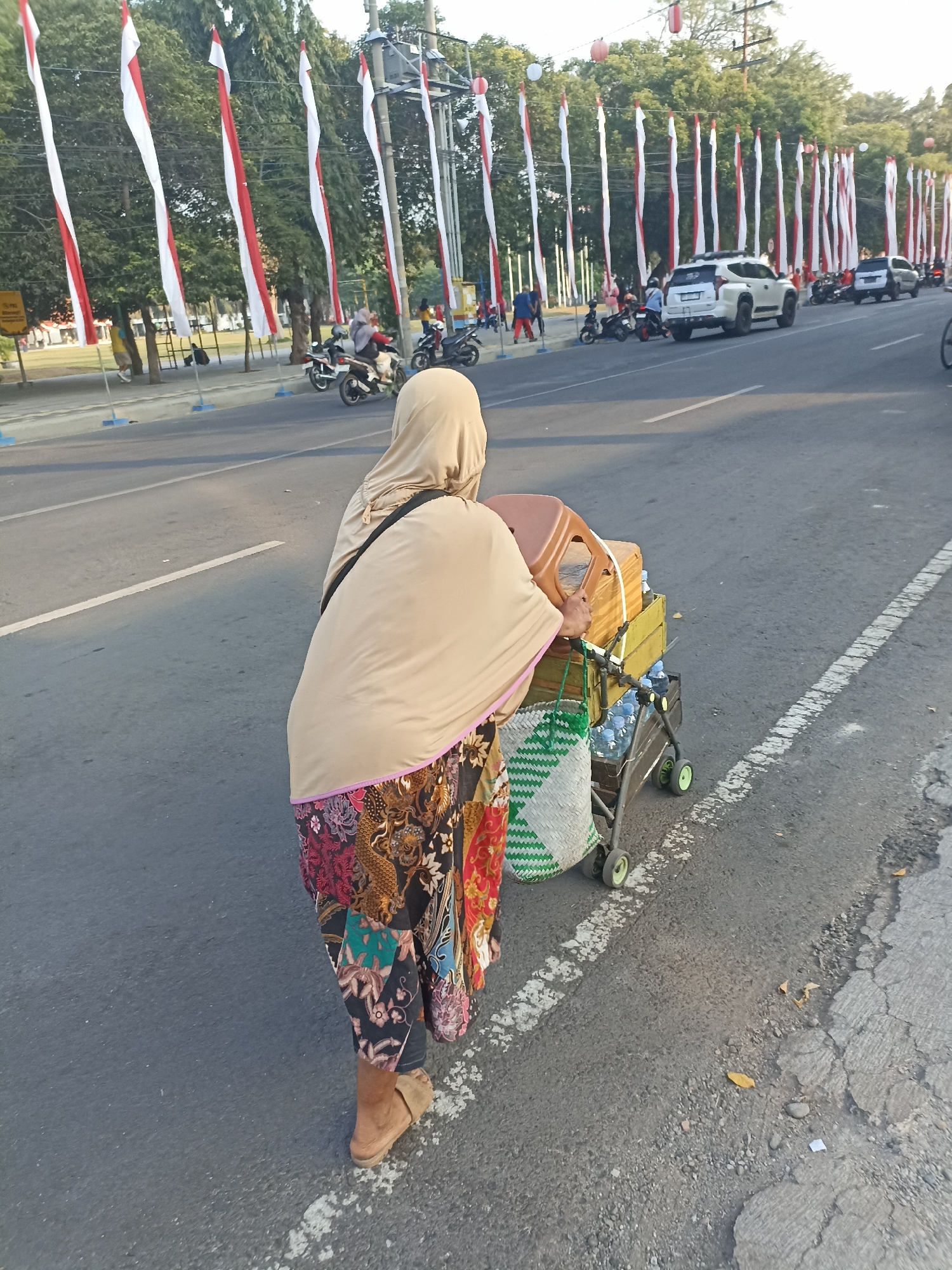 A person wearing a beige hijab and a colorful patterned skirt walks along a paved road, pushing a cart filled with bottled water, a wooden crate, and a plastic chair. The cart has green wheels. In the background, a row of Indonesian flags on poles lines the street, and several vehicles, including a white SUV and motorcycles, are visible. The scene appears to be in a city.