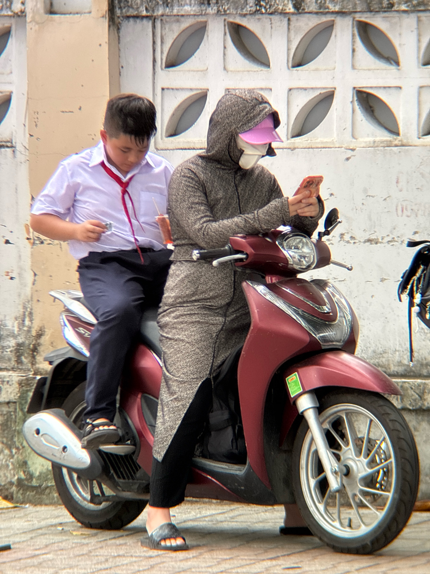 In Nha Trang, Vietnam, a young boy in a white school uniform and red tie sits on the back of a maroon scooter. He is looking down, holding a small object in his left hand and a straw in his right hand, with a cup of drink next to him. The person driving the scooter, likely an adult, is wearing a hooded gray jacket, a pink visor, and a white mask. They are holding a smartphone in their hands, looking at it. The scooter is parked on a paved surface, with a textured, off-white wall with decorative square cutouts in the background. The scene appears to be on a street or sidewalk.