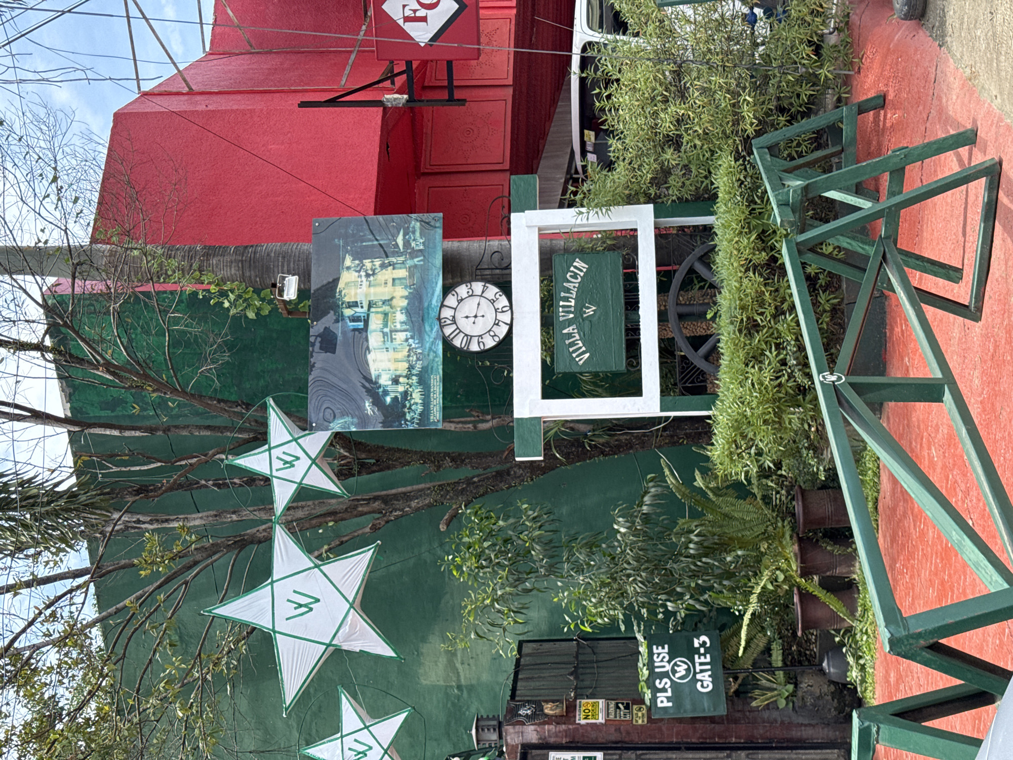 This image captures a vibrant exterior scene, likely an entrance or a section of a property, in Davao City, Philippines, under a clear daytime sky. The setting features a prominent red building or wall section in the background, topped by a white "F" logo. A large green wall serves as the primary backdrop for various decorative elements and signage.
Numerous plants and trees are visible, including a tree with bare branches draped with white star-shaped lanterns. These lanterns, known as parols, feature green outlines and a white "W" logo, suggesting a brand identity. Below them, a dark green sign mounted within a white rectangular frame clearly displays "VILLA VILLACIN W" in white text. To its left, a classic round clock with Roman numerals (visible from 12 to 7) and the "W" logo in its center tells a time of approximately 1:25. Above the clock, a large rectangular sign or artwork, possibly depicting a building, partially reveals the text "VILLA VILLACIN MANSION."
Closer to the foreground, a dark green sign with white text reads "PLS USE W GATE-3," indicating an entrance to the property. Nearby, several brown clay pots hold various ferns and leafy plants. Industrial-looking green metal barriers or structures are also visible on a red-colored ground surface, contrasting with the green foliage. A large, dark, spoked wheel, resembling a wagon wheel, is positioned behind the "VILLA VILLACIN" sign. The presence of the "W" logo across multiple items, along with "VILLA VILLACIN," strongly suggests this is a designated area of the Villa Villacin property. No people are visible, and the scene appears calm and well-maintained. A small "NO SMOKING" sign is also seen below the "PLS USE" sign.