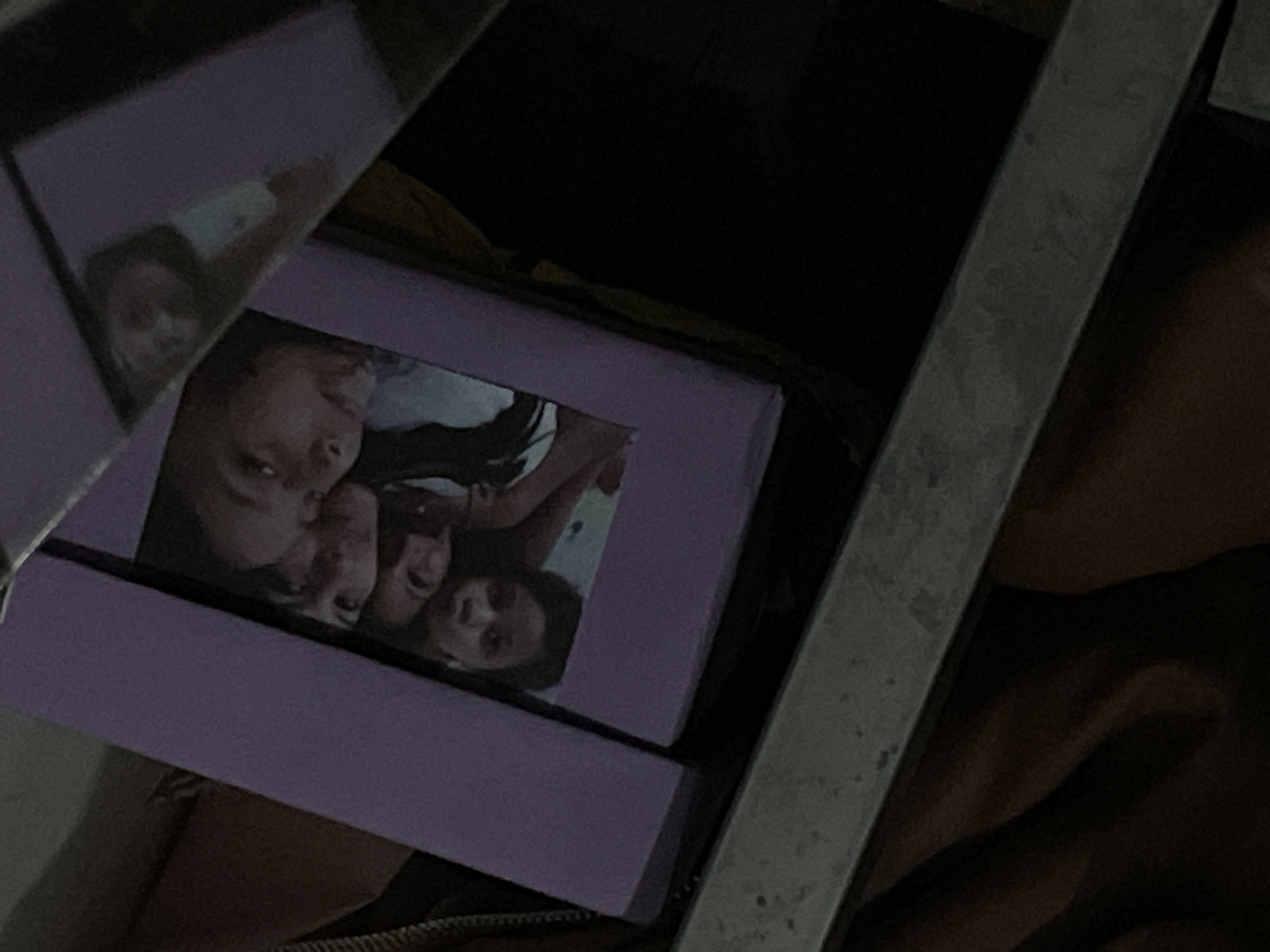 A close-up shot shows a purple box with a photo frame on its lid. The photo frame contains a picture of four young people, two in the foreground and two in the background. The box is resting on a dark surface, and a metal bar is visible on the left side of the image.