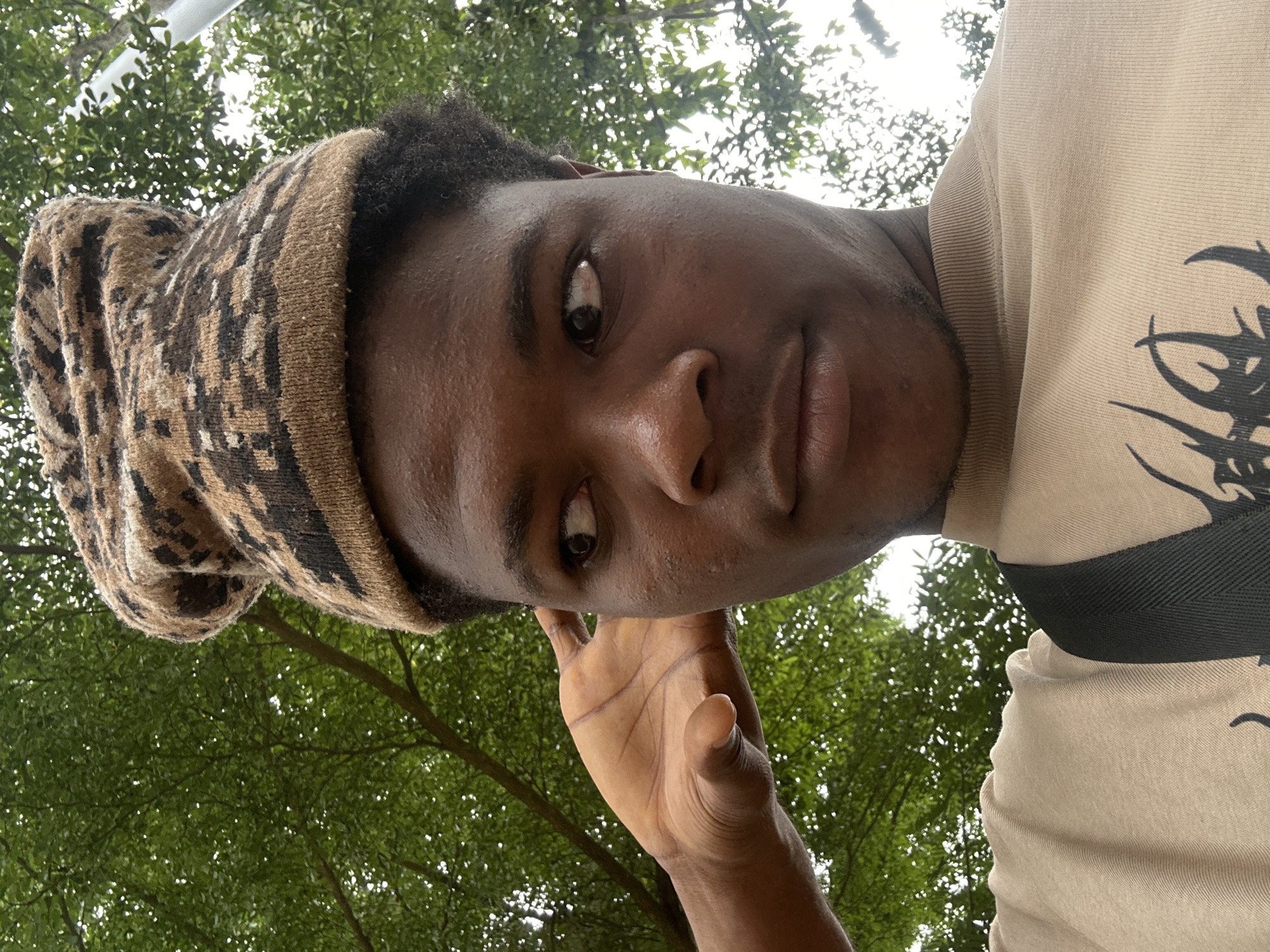 A low-angle, close-up selfie features a young Black male wearing a patterned knit beanie and a tan crewneck shirt. His right hand is raised to his ear, and he is looking to his left with his head tilted slightly. The background is dominated by the lush green foliage of trees, with some dappled sunlight filtering through. The overall impression is an outdoor setting, possibly a park or garden, on a bright day. The image is undated, but the general appearance of the subject and his attire suggests a contemporary setting. It's not possible to definitively pinpoint the location within Ihiagwa, Nigeria without further information, but the dense greenery is consistent with many areas in and around the city.