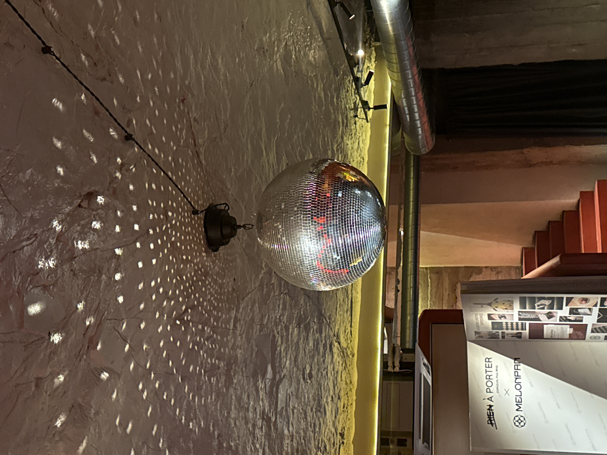 The image depicts the interior of a dimly lit venue with a textured, possibly concrete, ceiling. A large disco ball hangs from the ceiling, reflecting patterned light onto the textured surface. Below the ceiling, a bright yellow band of light is visible, possibly from strip lighting. Towards the right, a metal ventilation duct is prominent. In the foreground on the left, a display board or poster is visible, featuring text that appears to read "BIEN À PORTER" and "MELONPAN". To the right of this, a set of red stairs leads upwards. The overall ambiance suggests a club, bar, or event space. Without additional context or clearer signage, pinpointing the exact location within Yerevan, Armenia is not possible.