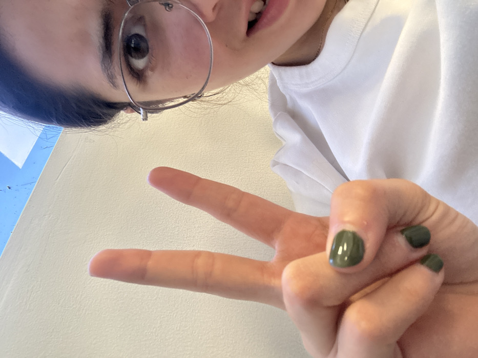 The selfie features a person with glasses making a peace sign. Their eye is visible through the lens. They wear a white shirt. The person's hand has dark green nail polish. The background appears to be a white or light-colored wall with a hint of blue at the upper left. The picture is taken from a slightly angled, high perspective.