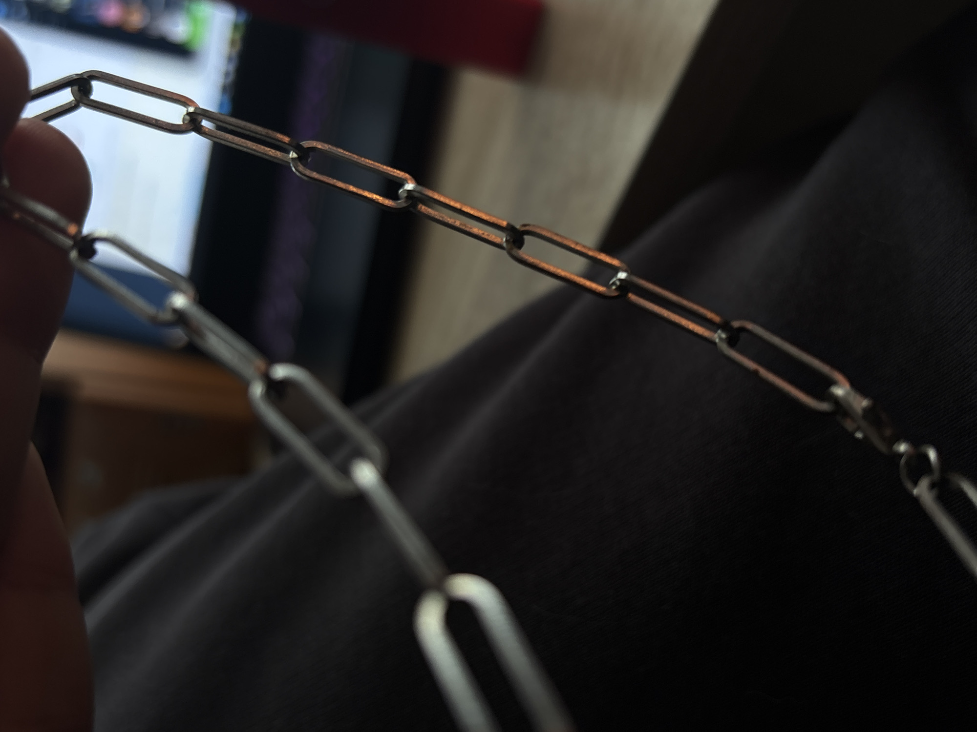 A close-up shot shows a metal chain, possibly made of stainless steel, with elongated oval links. The chain is held by a person's hand on the left side of the frame, with the fingers blurred due to the shallow depth of field. The chain extends diagonally across the image, draped over dark, textured fabric, likely clothing. The background is out of focus, with hints of a computer monitor displaying colorful graphics and a wooden surface suggesting an indoor setting. The lighting appears to be artificial, creating reflections on the metallic surface of the chain and casting subtle shadows. No specific time of day or weather conditions are discernible. The overall mood is neutral, focusing on the detail and texture of the object. No visible text is present in the image. The location context of Saint Petersburg, Russia, provides a general geographic setting for the scene.