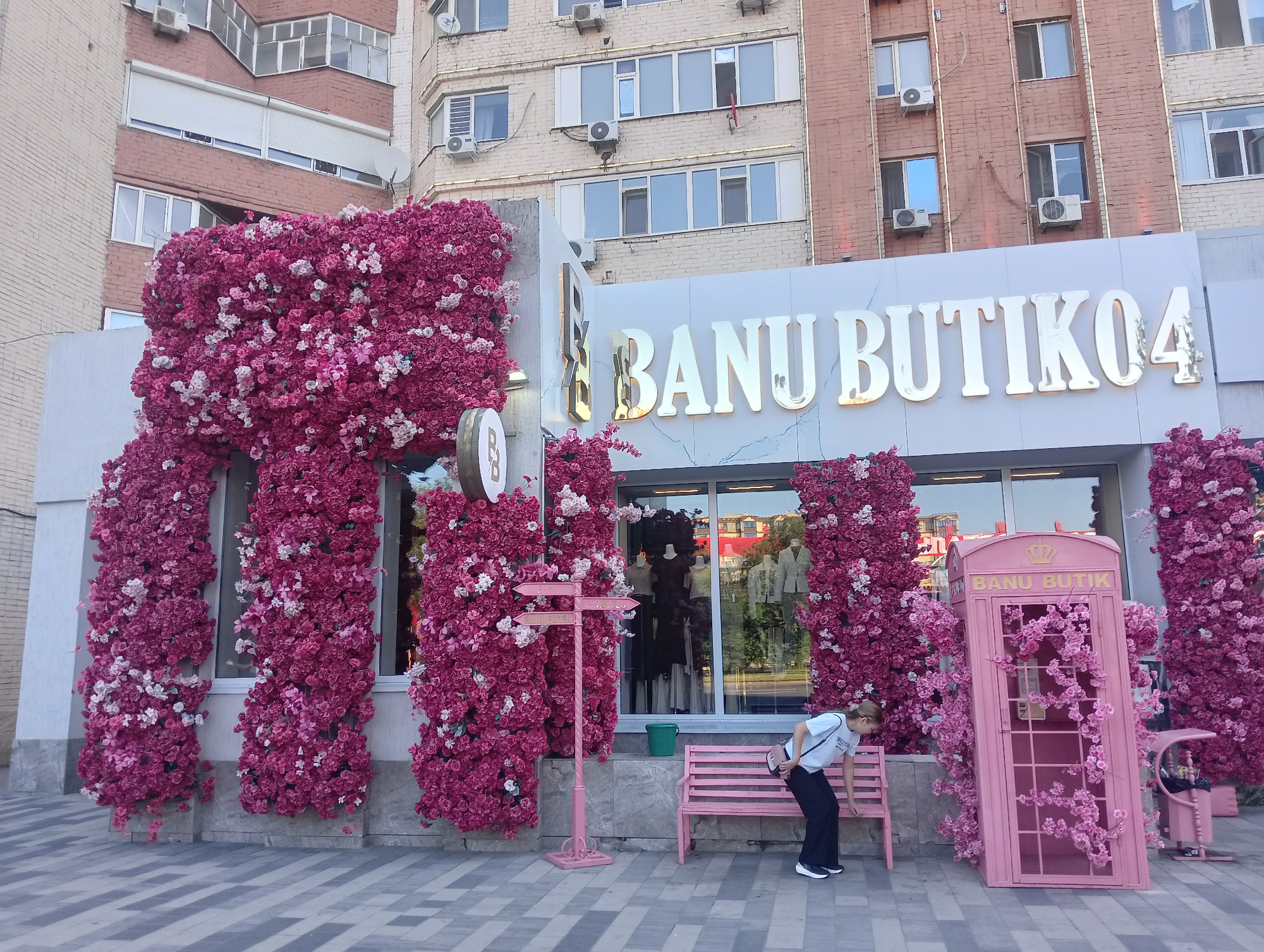 In Aktobe, Kazakhstan, a shopfront is elaborately decorated with a profusion of pink and white artificial flowers. The flowers frame the entrance and spill onto the facade of the building. Above the entrance, a prominent sign reads "BANU BUTIK04" in large, gold, serif letters against a white marble-patterned background. To the right of the entrance, a pink, vintage-style telephone booth, also adorned with pink flowers, stands as a decorative element. In front of the shop, a young person wearing a white t-shirt and black pants is leaning on a pink bench. The ground is paved with a geometric pattern of grey and light grey tiles. The background shows a multi-story building with several windows and air conditioning units.