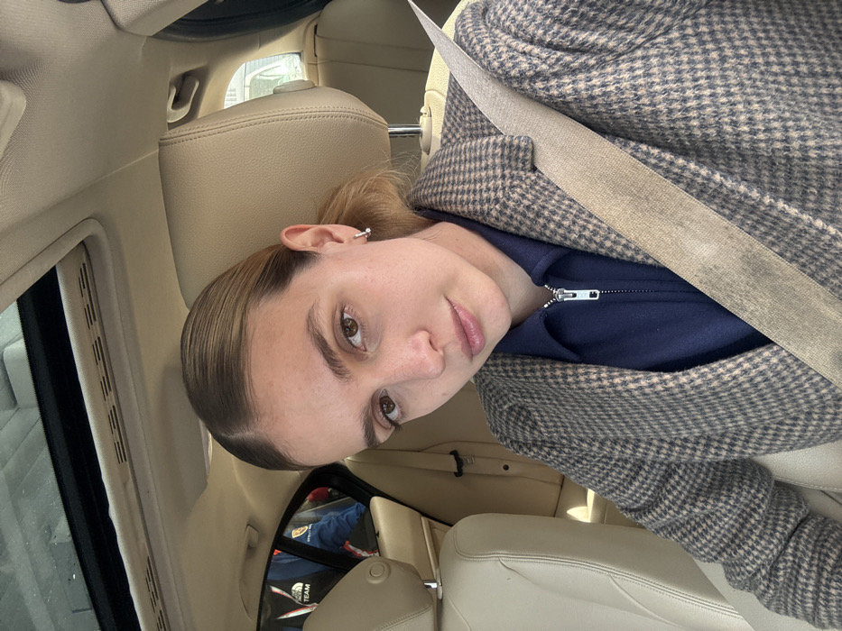 A young adult female is depicted from the torso up, seated in the front passenger seat of a car. She has light brown hair neatly pulled back from her face with a middle part and is looking directly at the camera. She wears a dark blue collared shirt with a silver zipper, layered under a brown and beige houndstooth or plaid pattern blazer. A light tan seatbelt diagonally crosses her chest. A small silver hoop earring is visible on her left ear. The car's interior features beige leather upholstery, including the seats and headliner, and a sunroof is visible above. Through the rear passenger window, a portion of another individual wearing a blue jacket with a "THE NORTH FACE TEAM" logo is visible. The scene is located in the city of Tbilisi, Georgia.