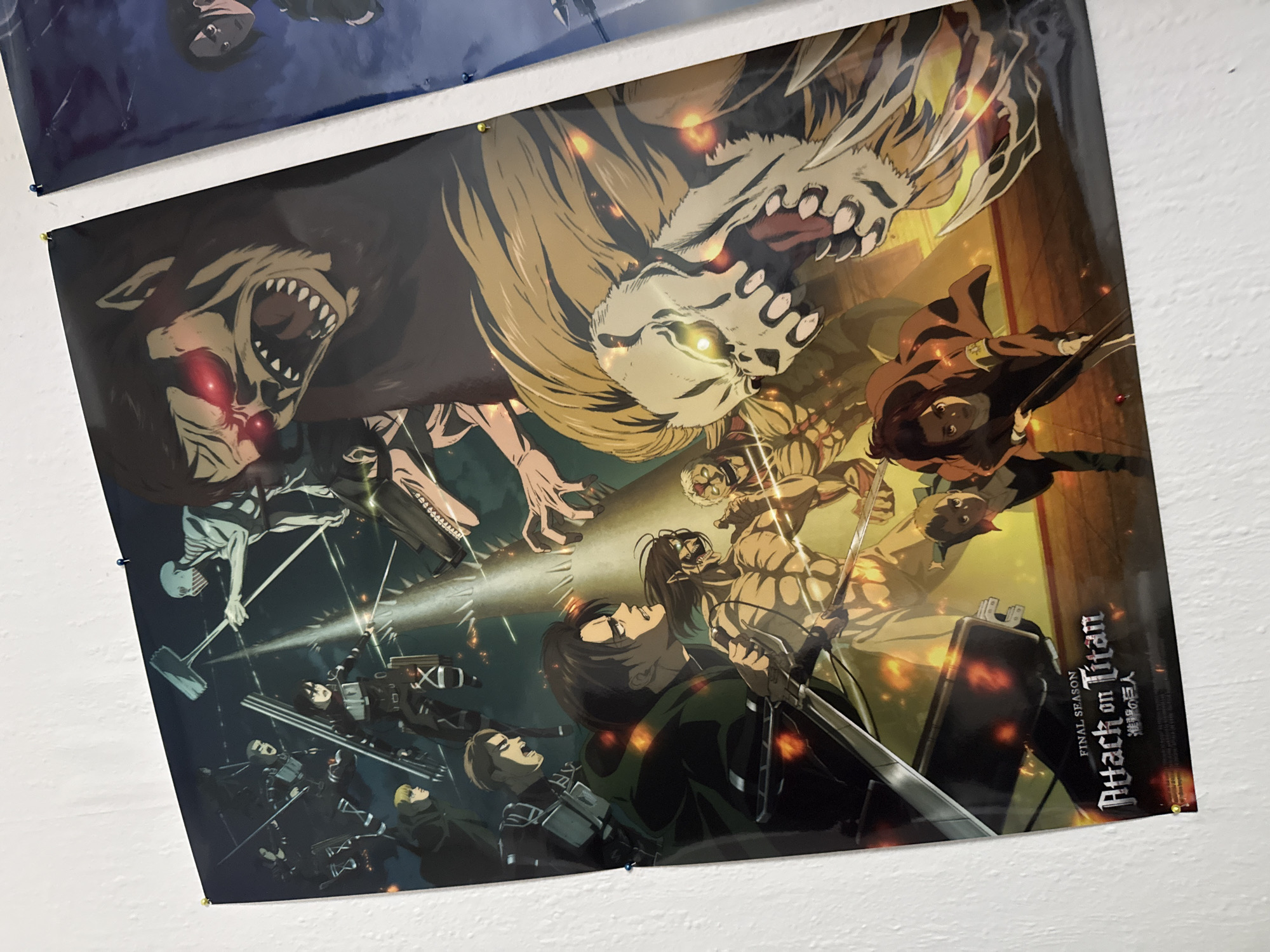 A poster for the anime series "Attack on Titan" is displayed on a white wall. The poster features several characters from the series, including Eren Yeager in his Titan form, Levi Ackerman, and Mikasa Ackerman, engaged in combat with various Titans. The scene is dynamic and action-packed, with Titans roaring and characters wielding swords. The poster is pinned to the wall with yellow and blue pins, with one blue pin visible on the right edge of the poster. Another poster, partially visible on the right, appears to be a similar anime artwork. The environment is a plain white wall, suggesting an indoor setting. This scene is located in the town of Bad Sachsa, Germany.